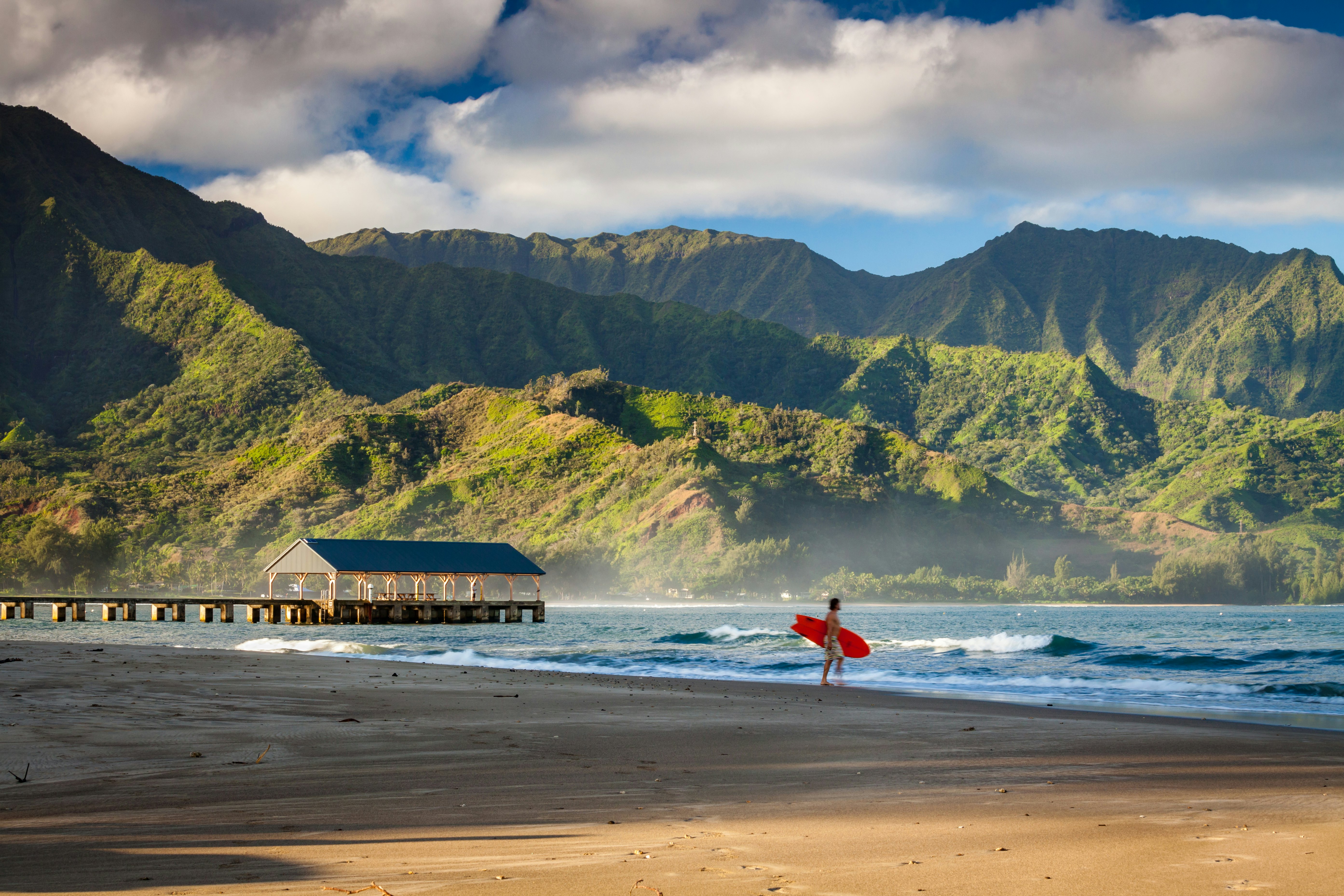 A surfer heads out into the waves at a beach surrounded by high volcanic hills