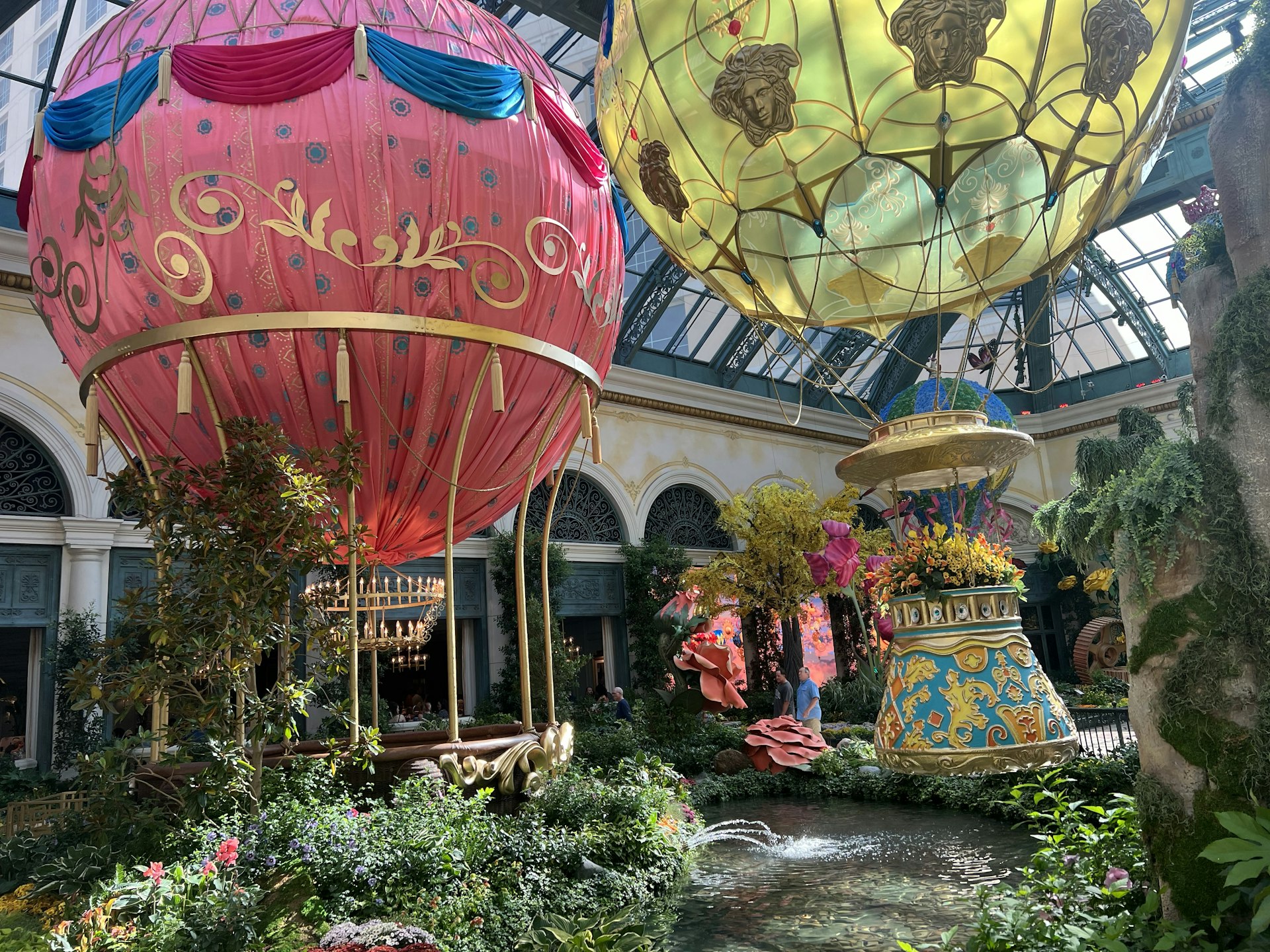 Outside view of the Garden Table (under the red hot-air ballon) located in the Bellagio Conservancy and Botanical Garden. There's a second yellow hot-air ballon suspended above the garden. There are green plants, flowers and small pond in the foreground.
