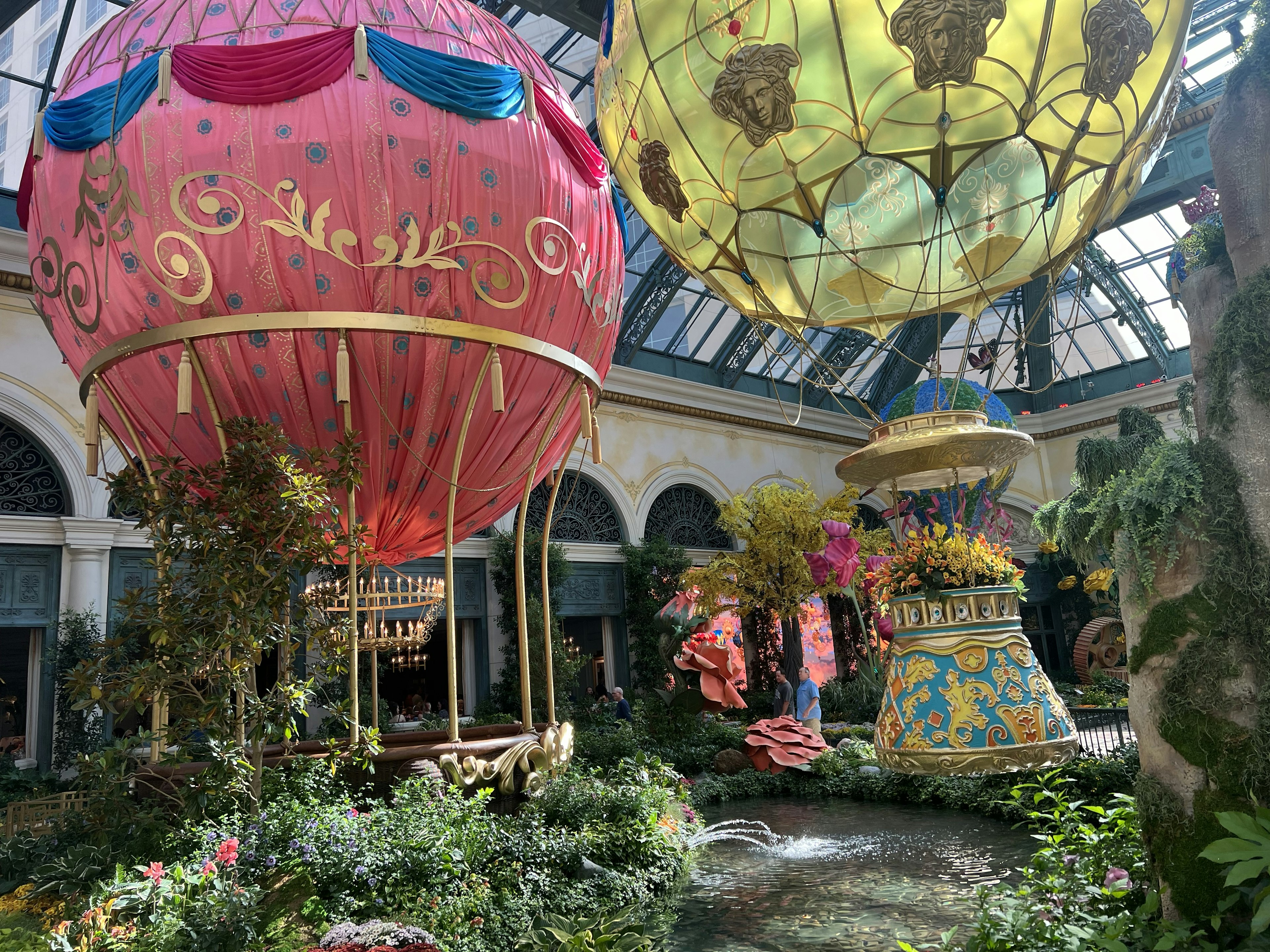 Outside view of the Garden Table (under the red hot-air ballon) located in the Bellagio Conservancy and Botanical Garden. There's a second yellow hot-air ballon suspended above the garden. There are green plants, flowers and small pond in the foreground.