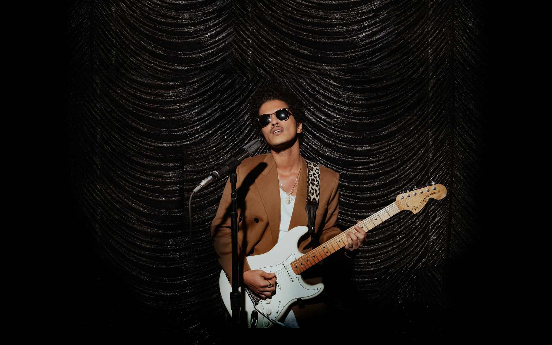 Bruno Mars stands in front of a microphone, holding a creme and white guitar. There's a black curtain in the background. 
