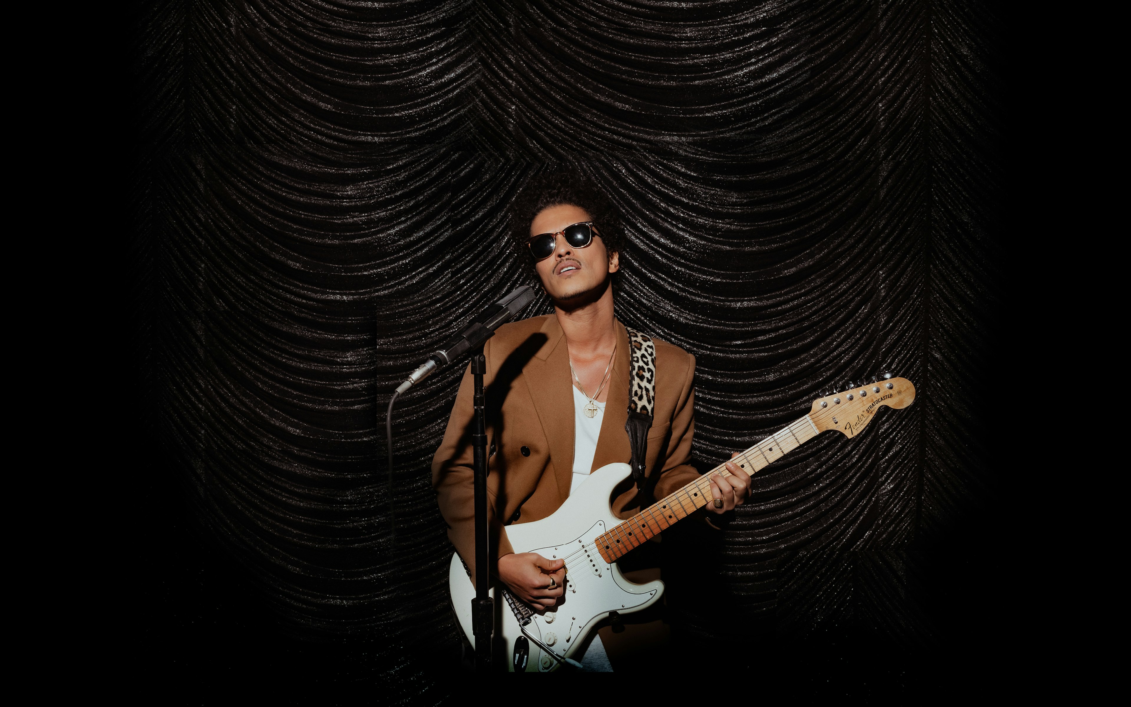 Bruno Mars stands in front of a microphone, holding a creme and white guitar. There's a black curtain in the background.