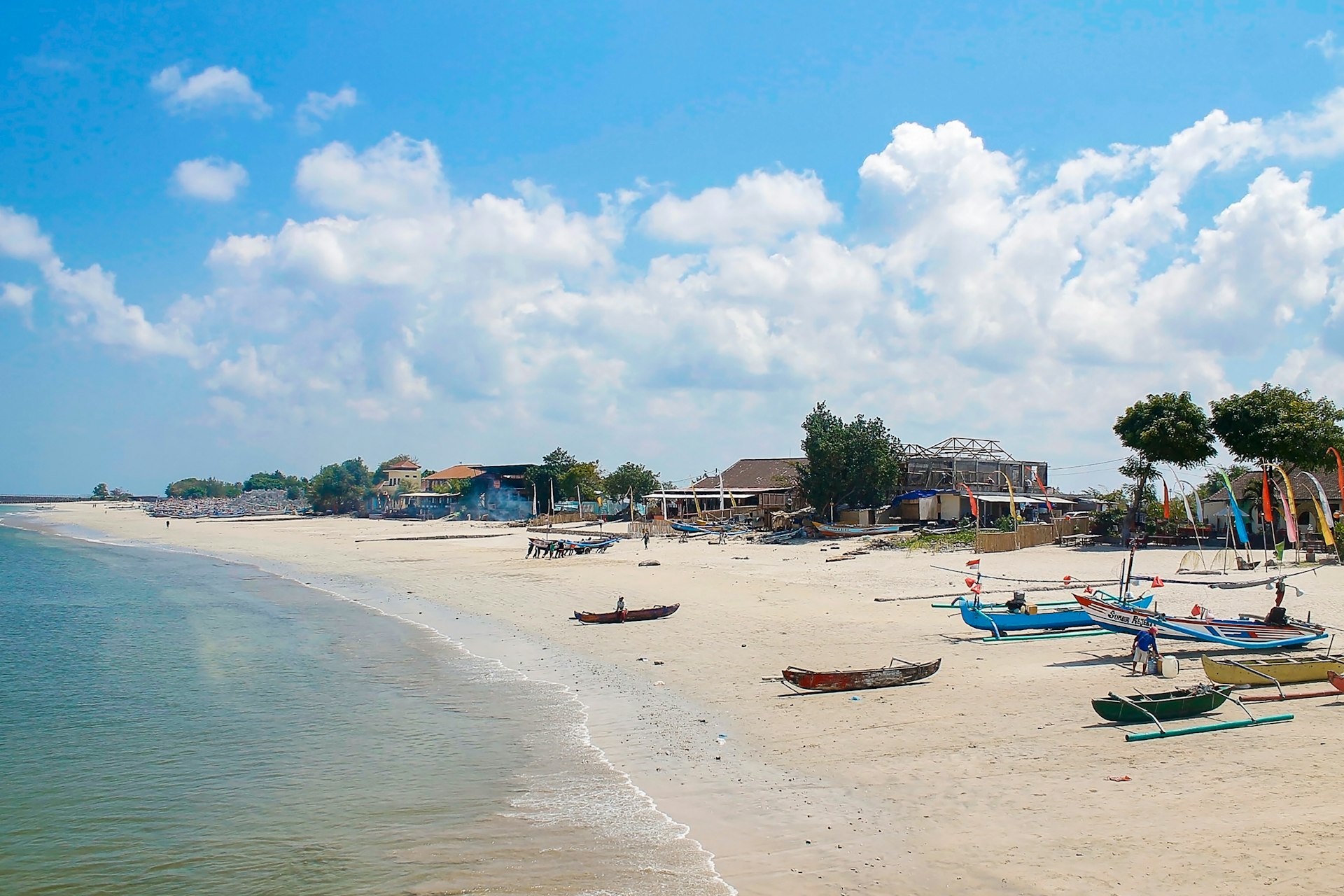 Small fishing boats are lined up on a sandy beach, with simple buildings beyond.