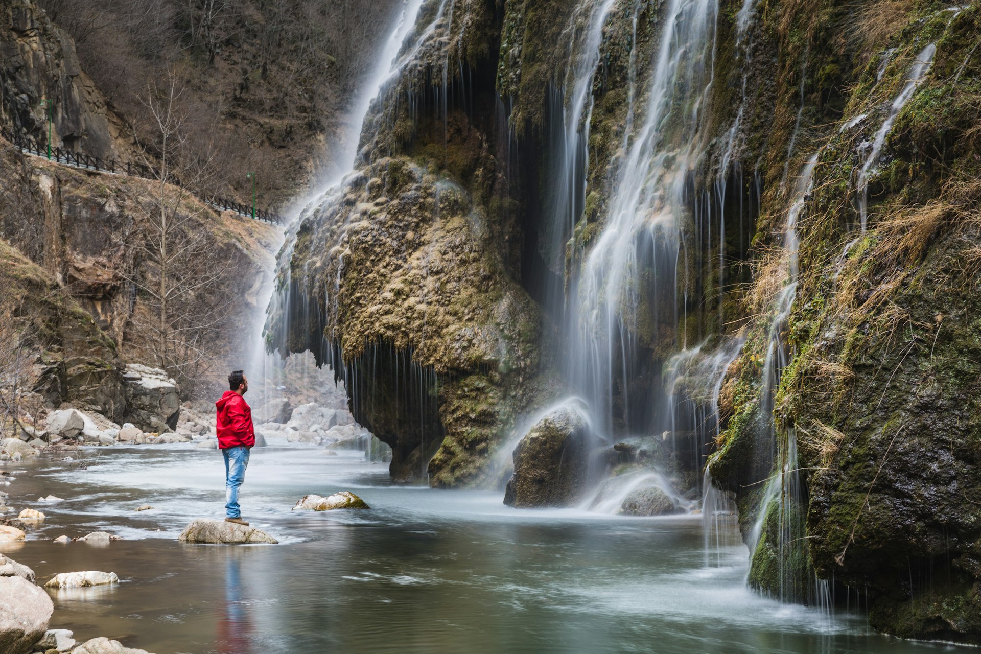 A man in red wet-weather gear and hiking boots stands on a rock in a pool watching a waterfall