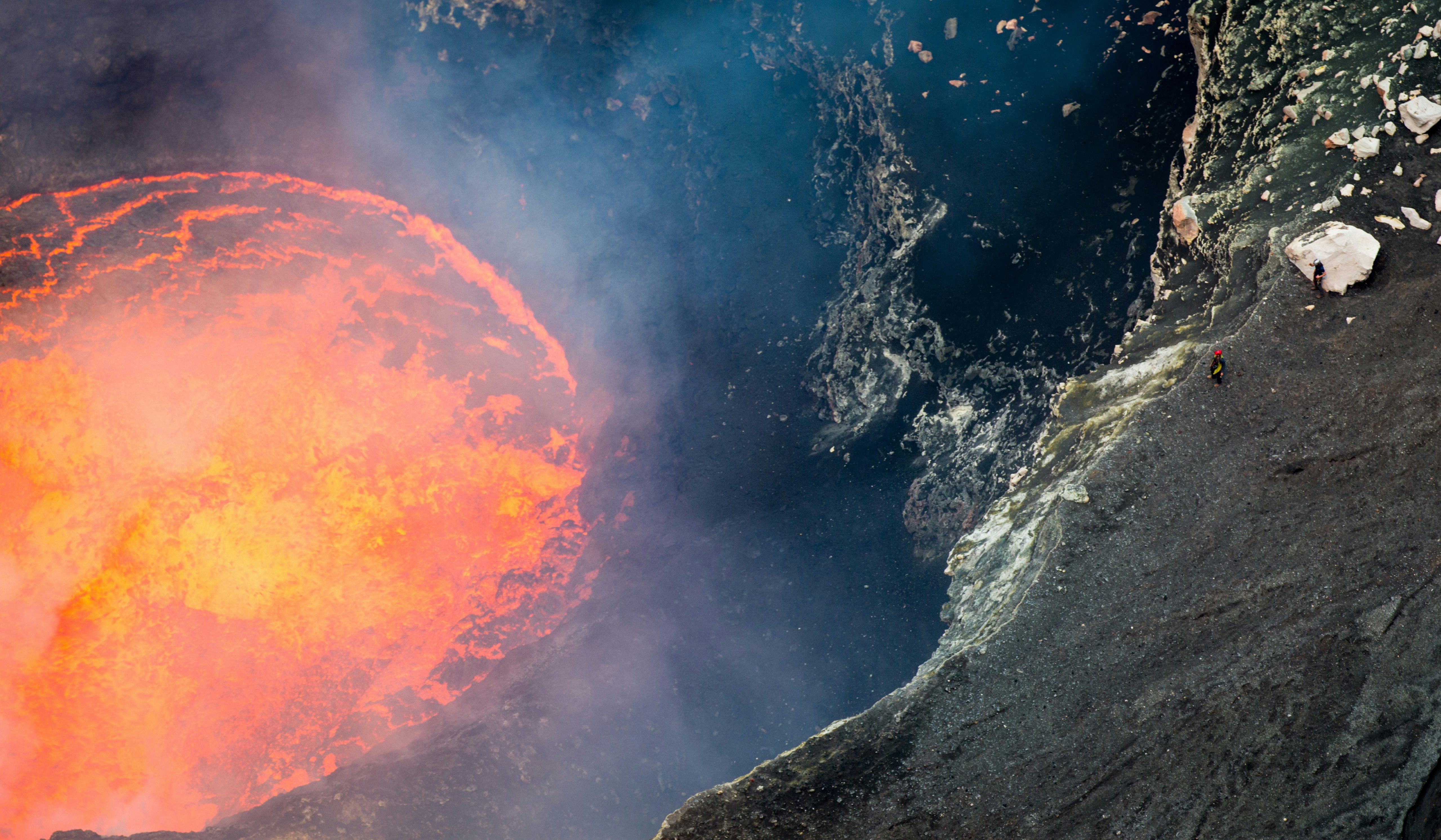 An orange lake of lava with two hikers near the rim gazing downwards