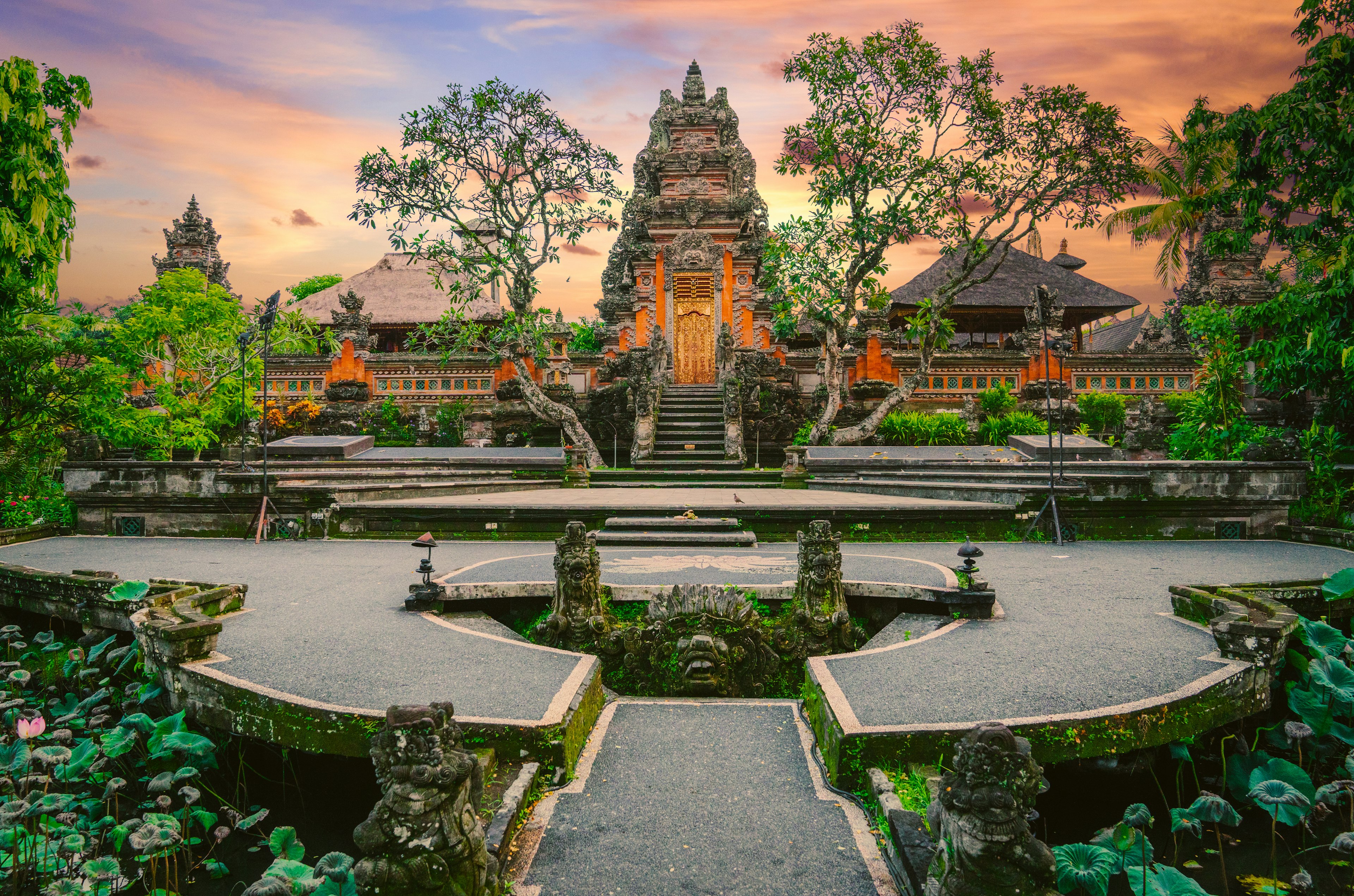 A pathway between two lotus-flower ponds leads towards a pale red colored temple