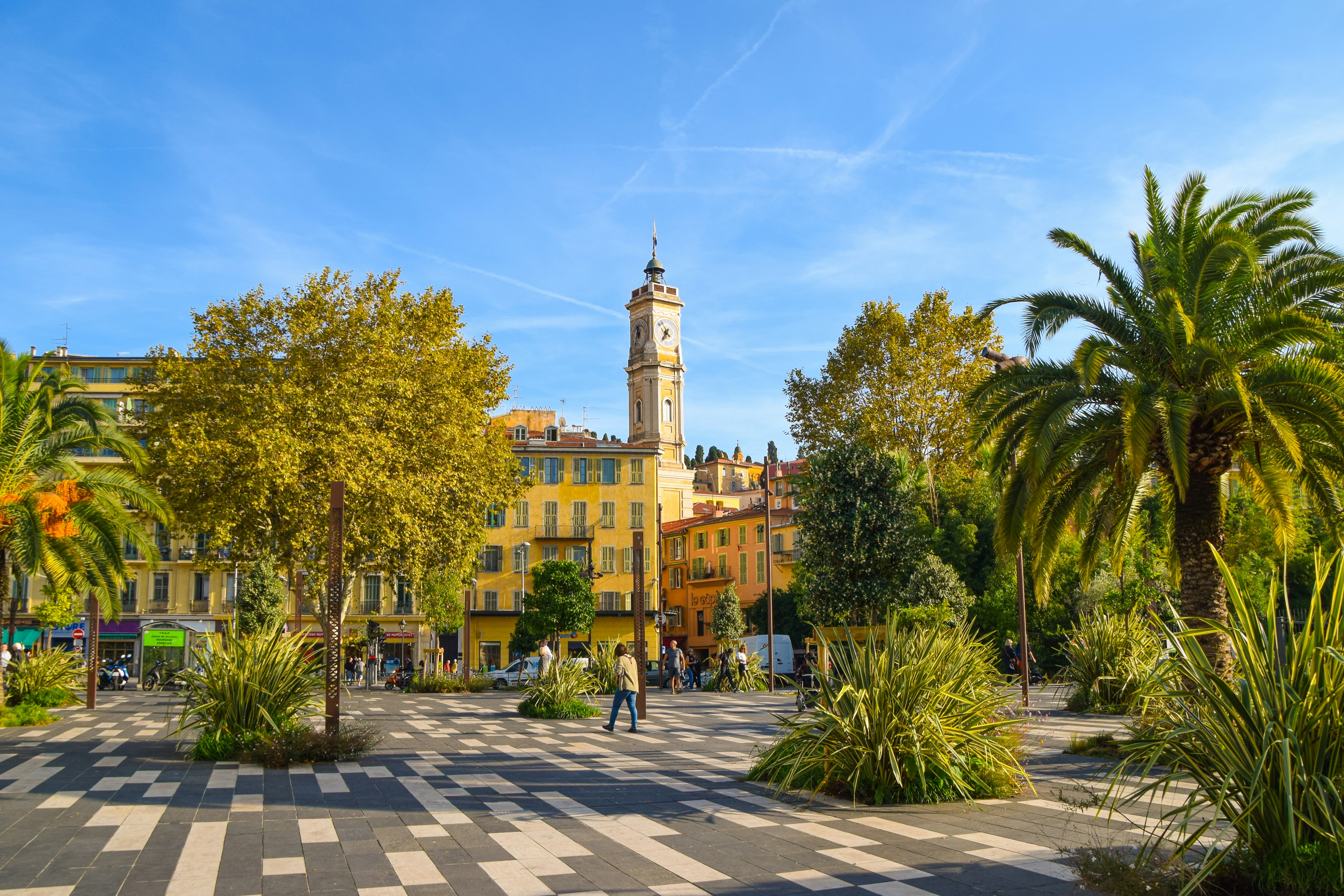 Several palm trees of various sizes are in the foreground at the Promenade du Paillon in the Old Town of Nice, France, with the St Francois tower in the background.