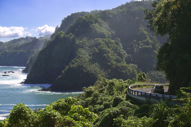 A car drives around a bend that is hugging a cliffside covered in green foliage