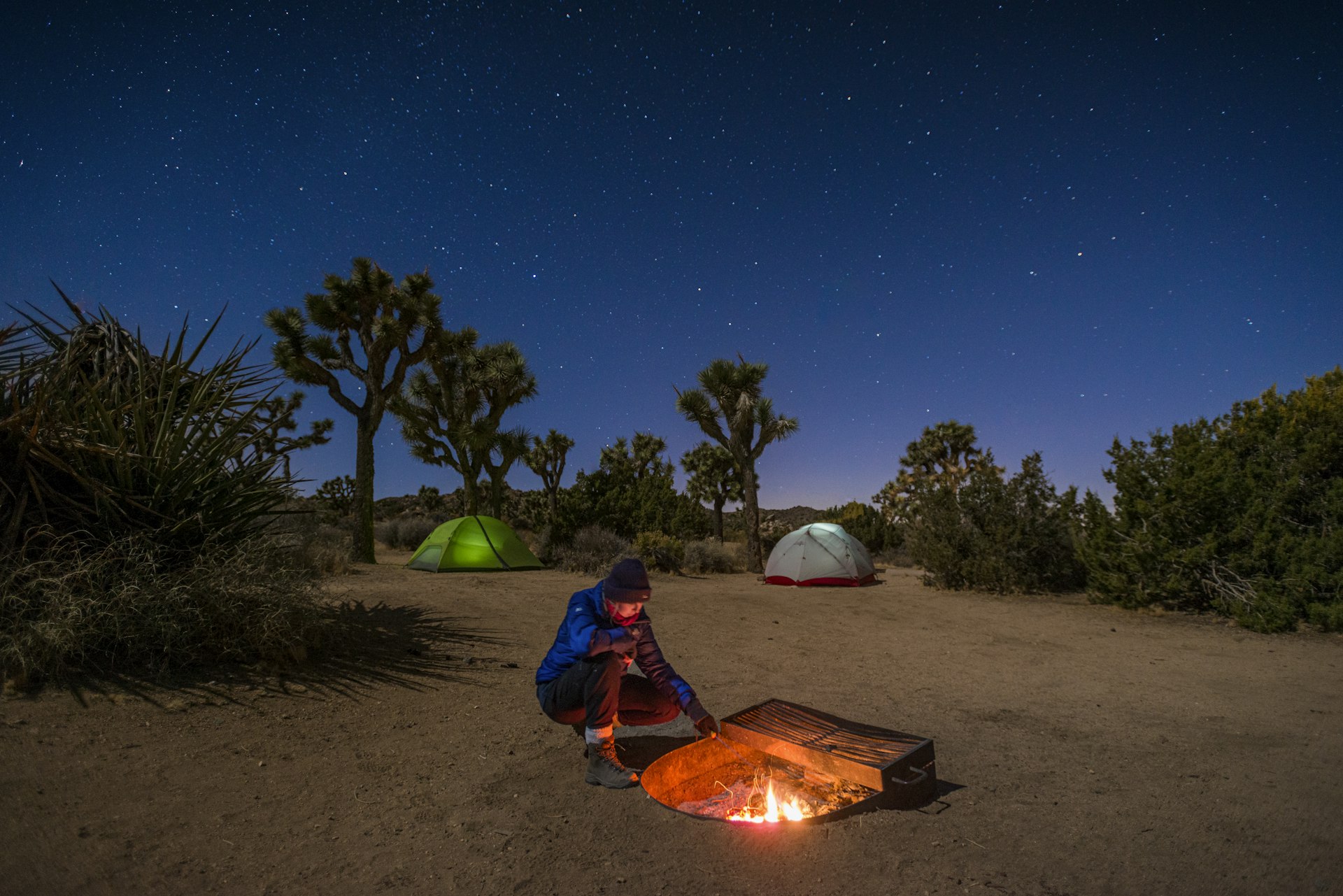 A woman stokes a camp fire near her tent at night in Joshua Tree National Park, California, USA