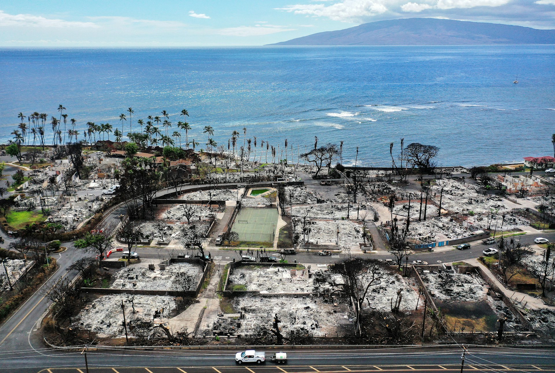 In an aerial view, a recovery vehicle drives past burned structures and cars two months after a devastating wildfire on October 9, 2023 in Lahaina, Maui, Hawaii