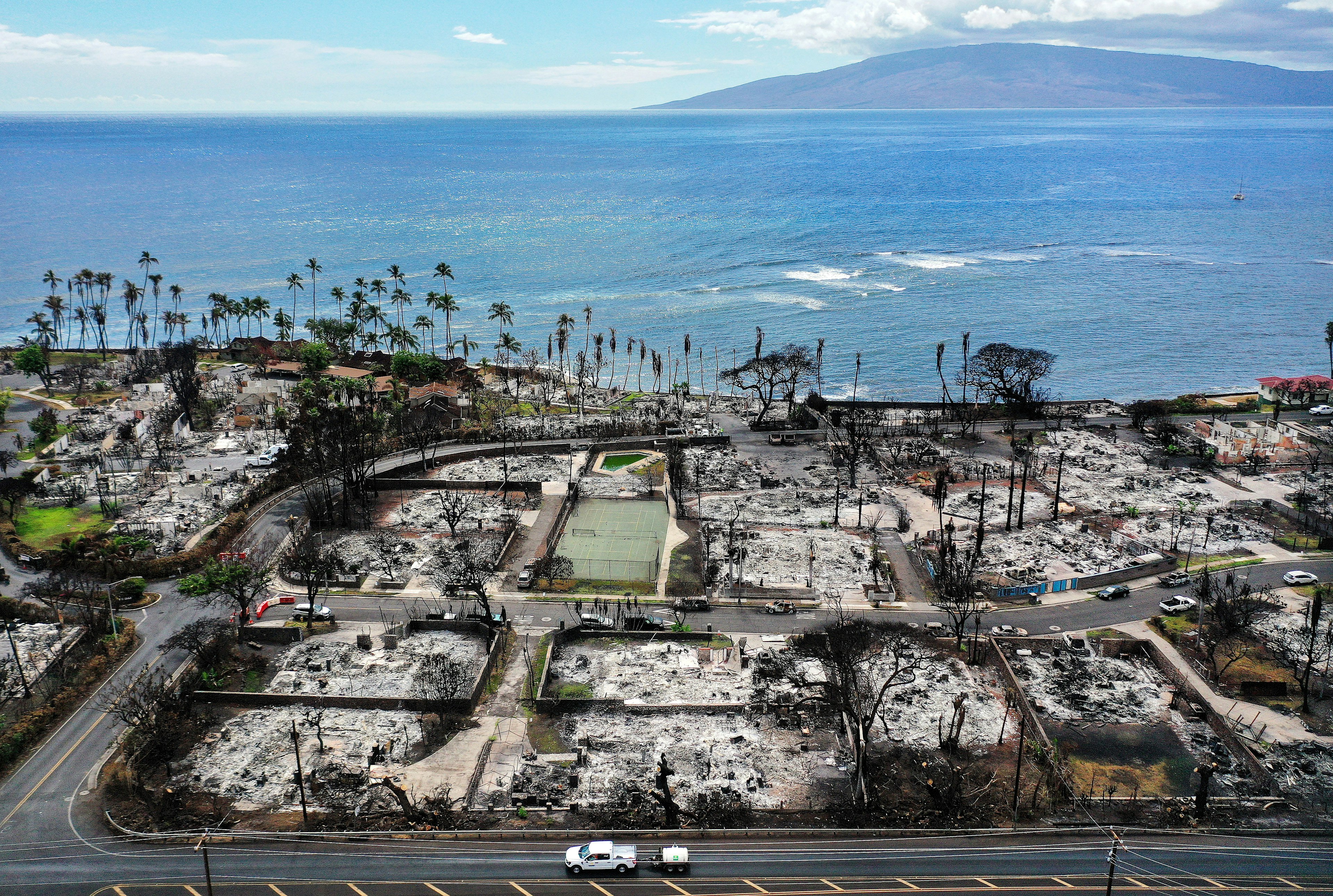 In an aerial view, a recovery vehicle drives past burned structures and cars two months after a devastating wildfire on October 9, 2023 in Lahaina, Maui, Hawaii