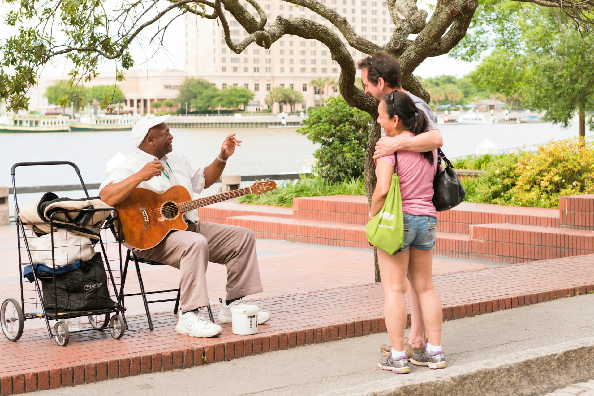 A couple have their arms around each other while they stand and chat to a street performer next to a river