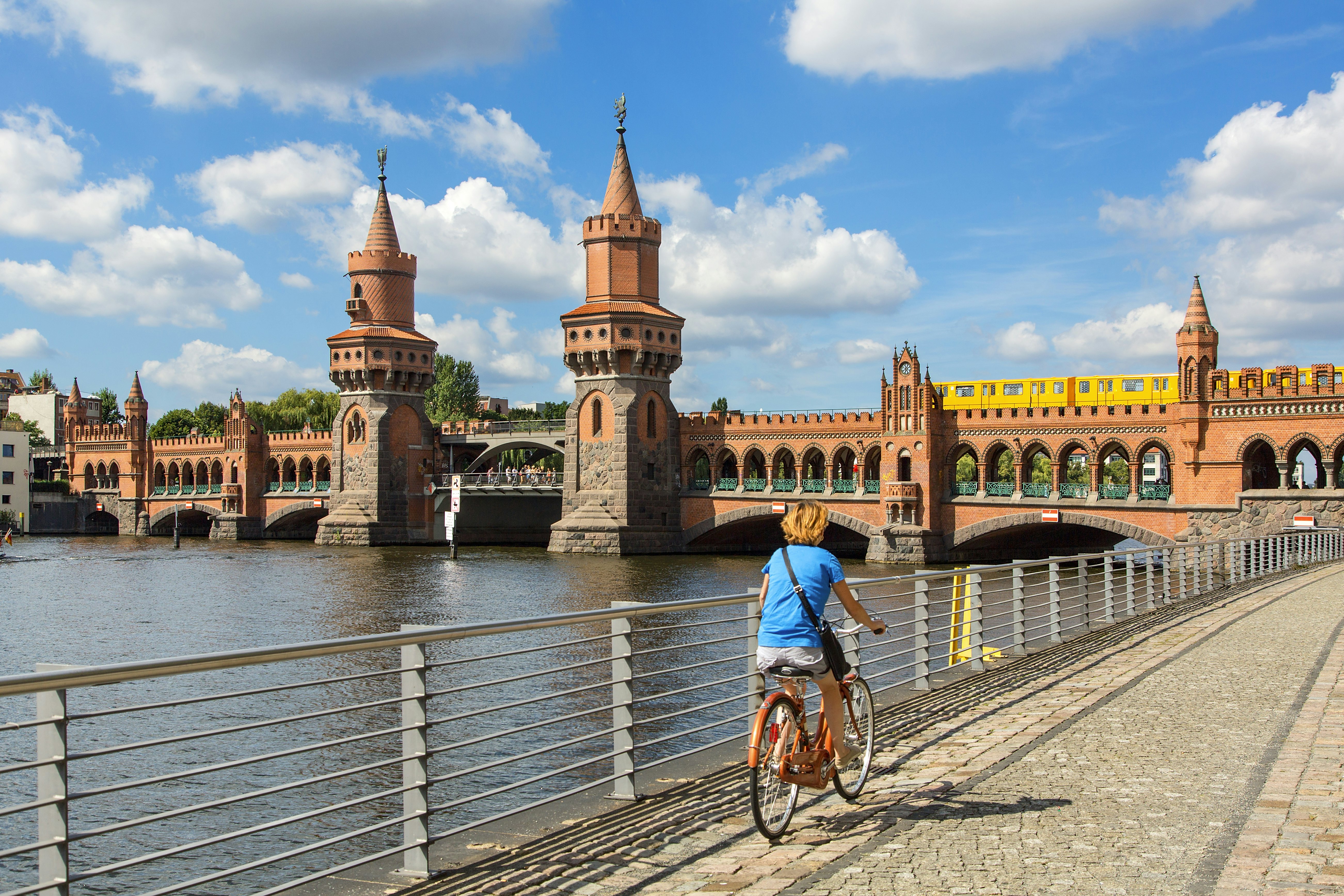 A cyclist rides a bike along a riverside path towards a bridge where a train is crossing