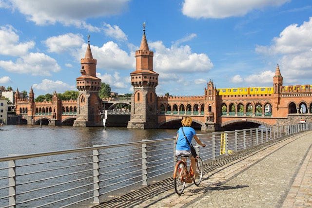 A cyclist rides a bike along a riverside path towards a bridge where a train is crossing