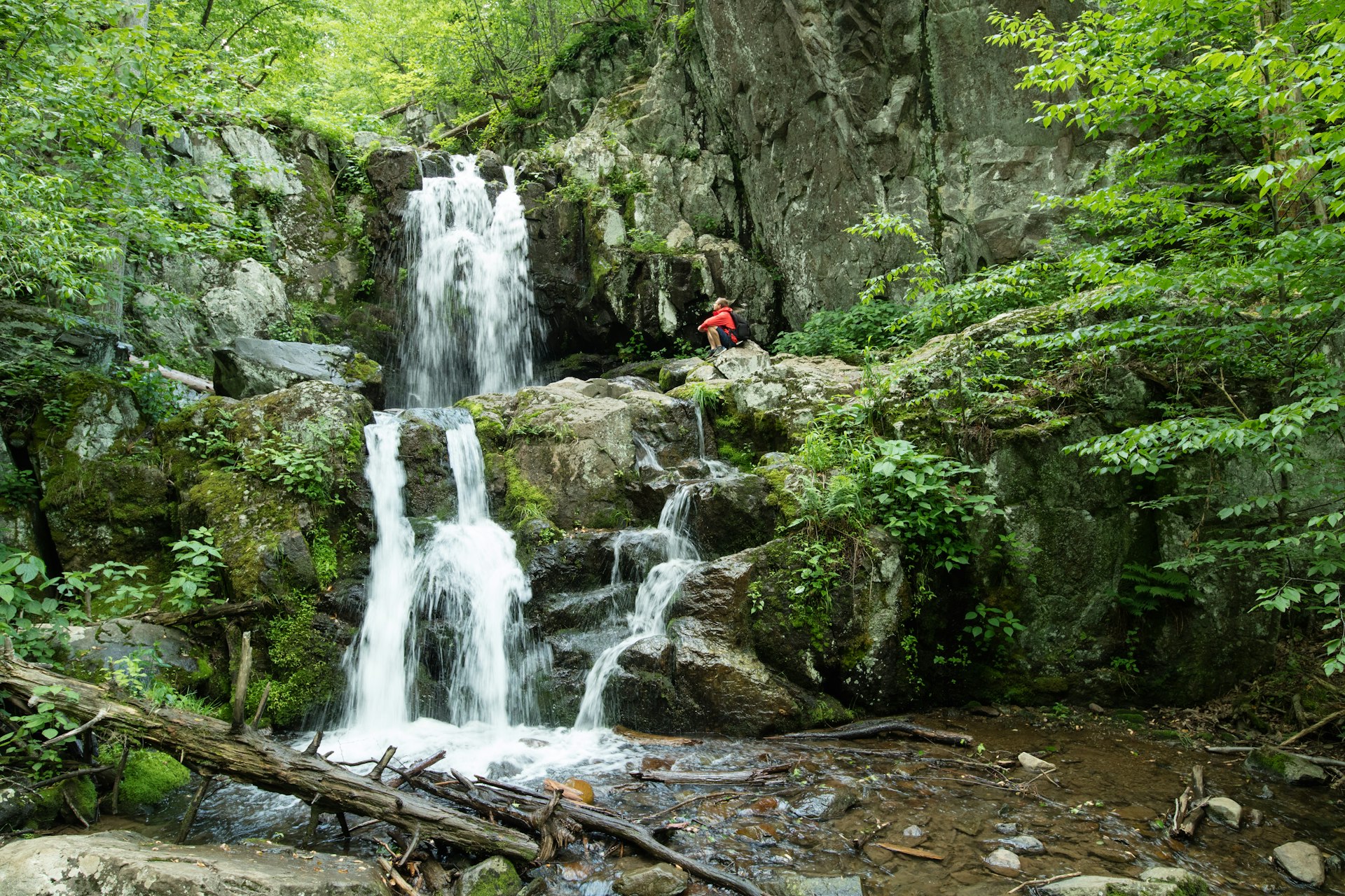 A hiker sits on rocks near a cascading waterfall