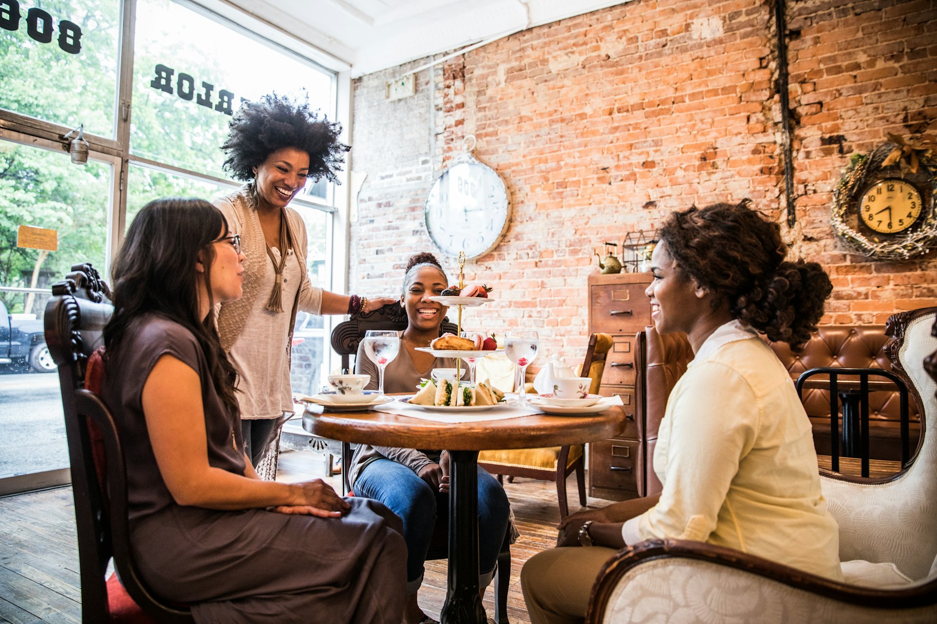 Three women are sitting around a table in a tea parlor, while another woman stands next to the table, talking to them