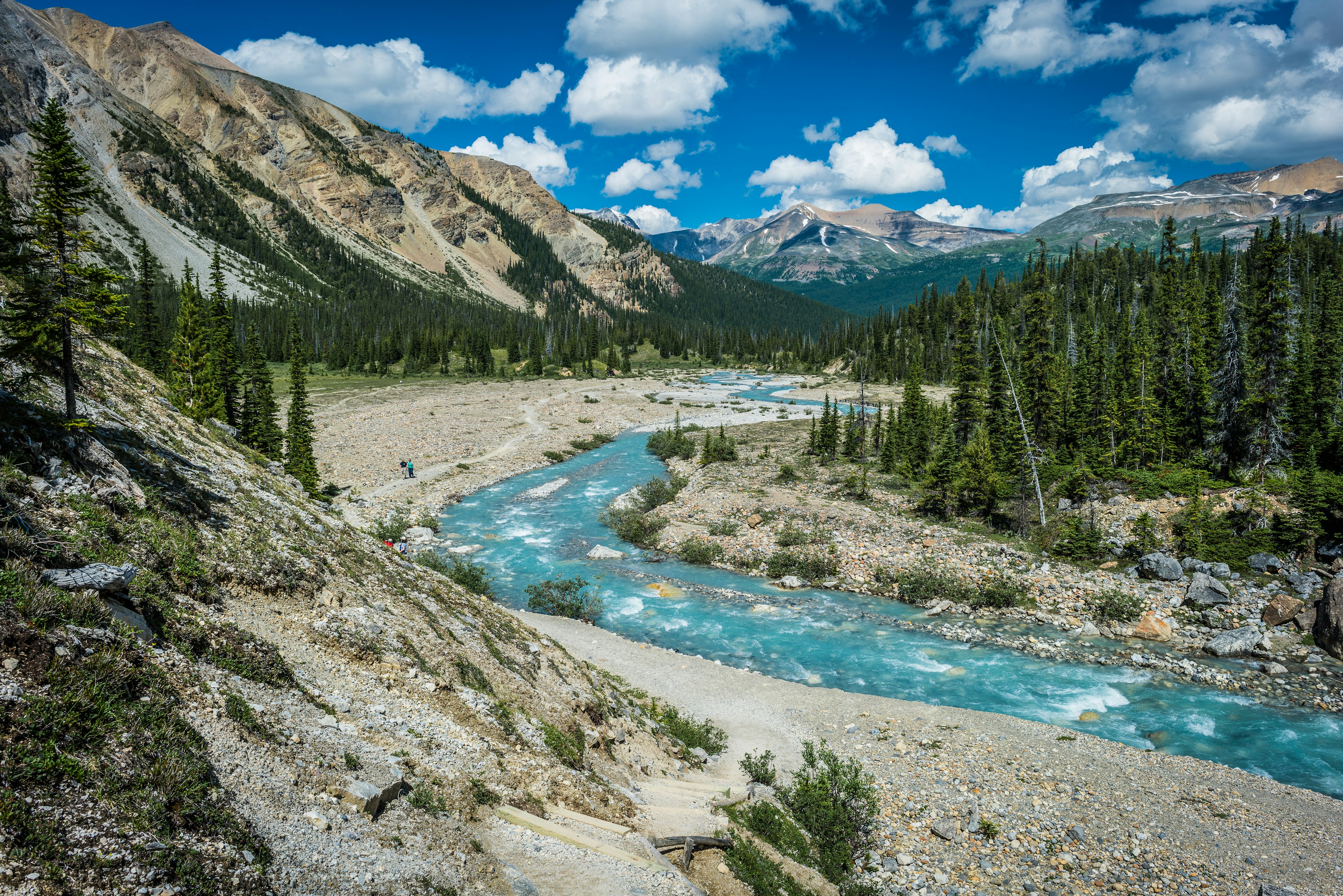 A rushing river runs through a valley; some people follow a hiking trail nearby