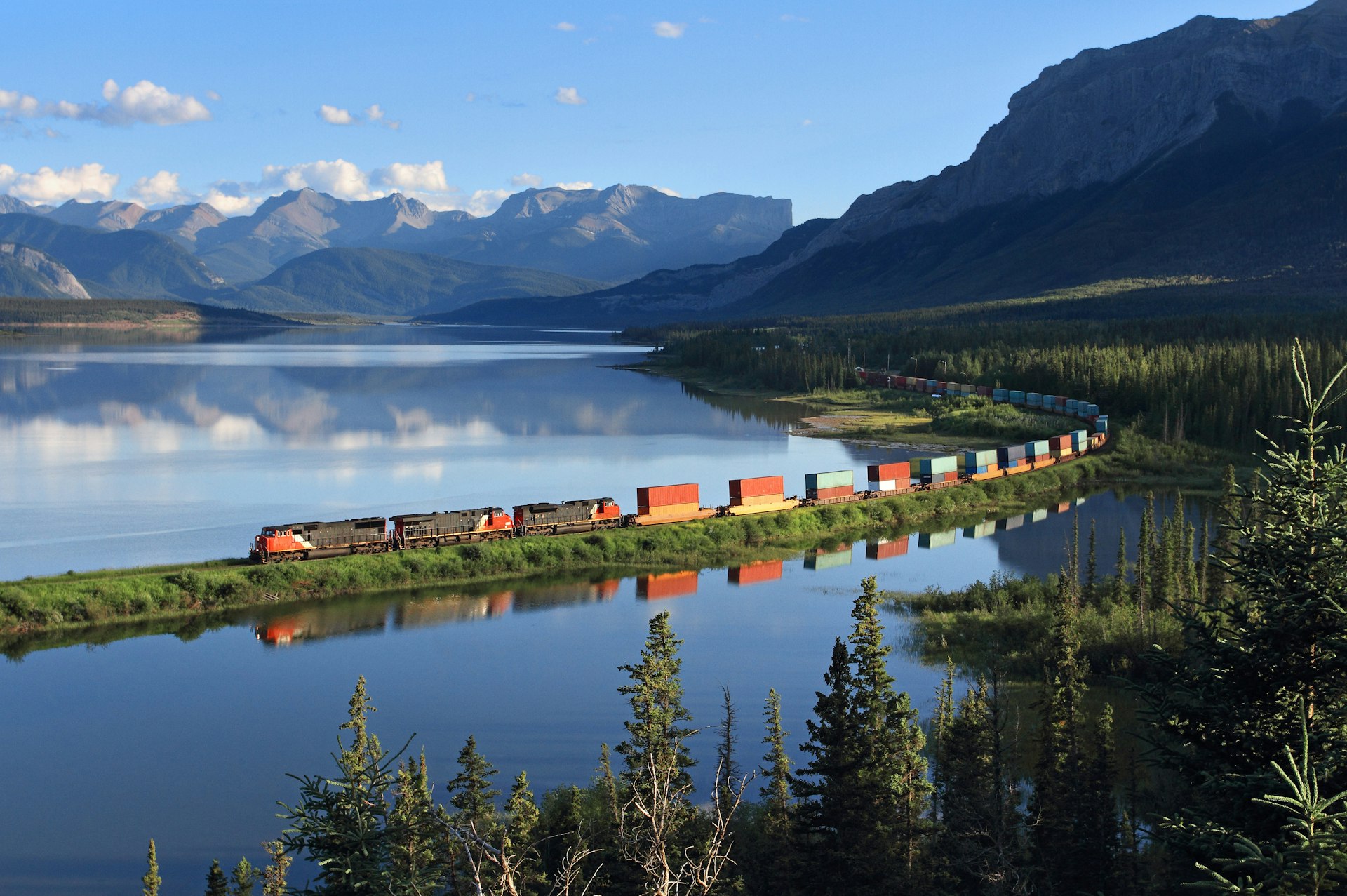 A long freight train follows the tracks through an incredibly scenic lakeside and mountain location