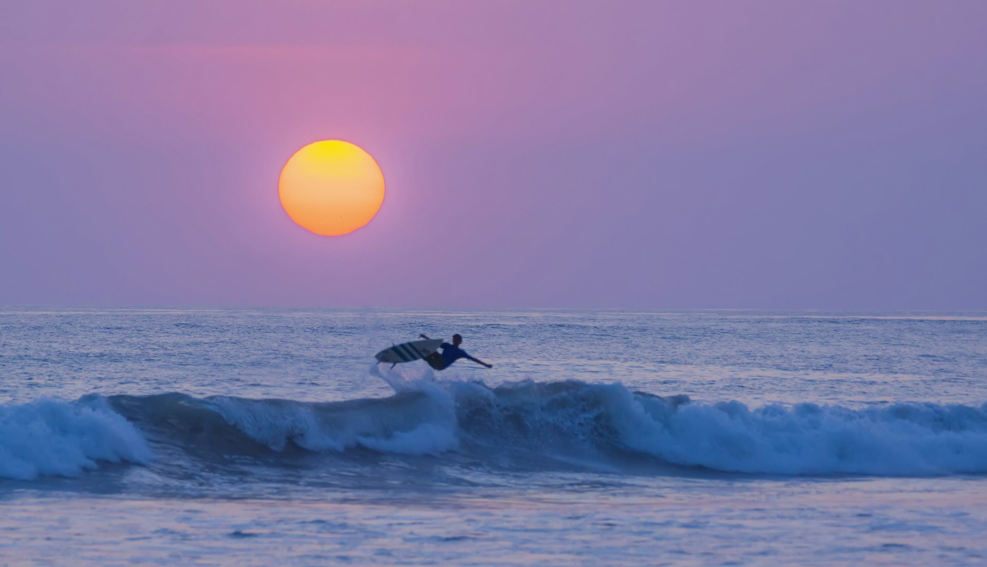 A surfer hits the top of a wave. The orange sun hangs low in the sky