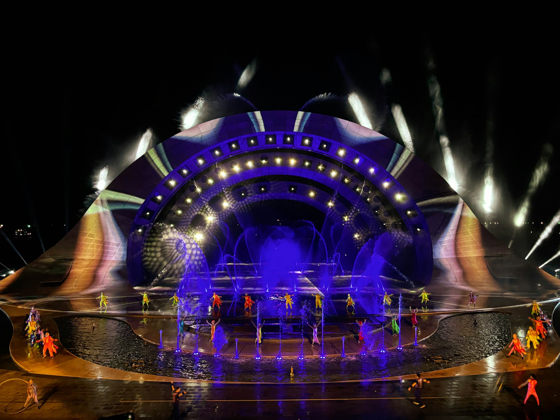 Many dancers are moving on a vast stage with fountains, illuminated by a complex lightshow