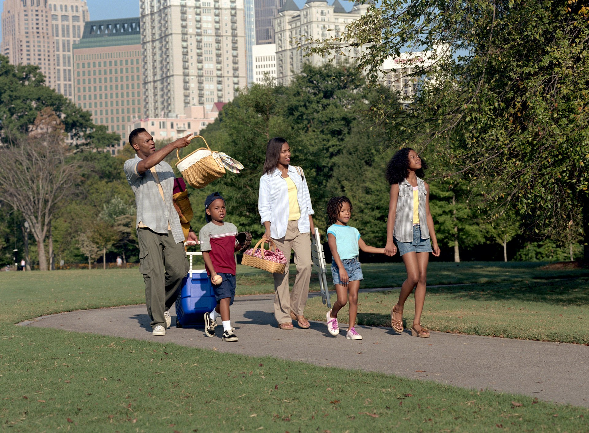 A family of five walking along a path in Piedmont Park, Atlanta, with picnic baskets and a cooler on wheels.