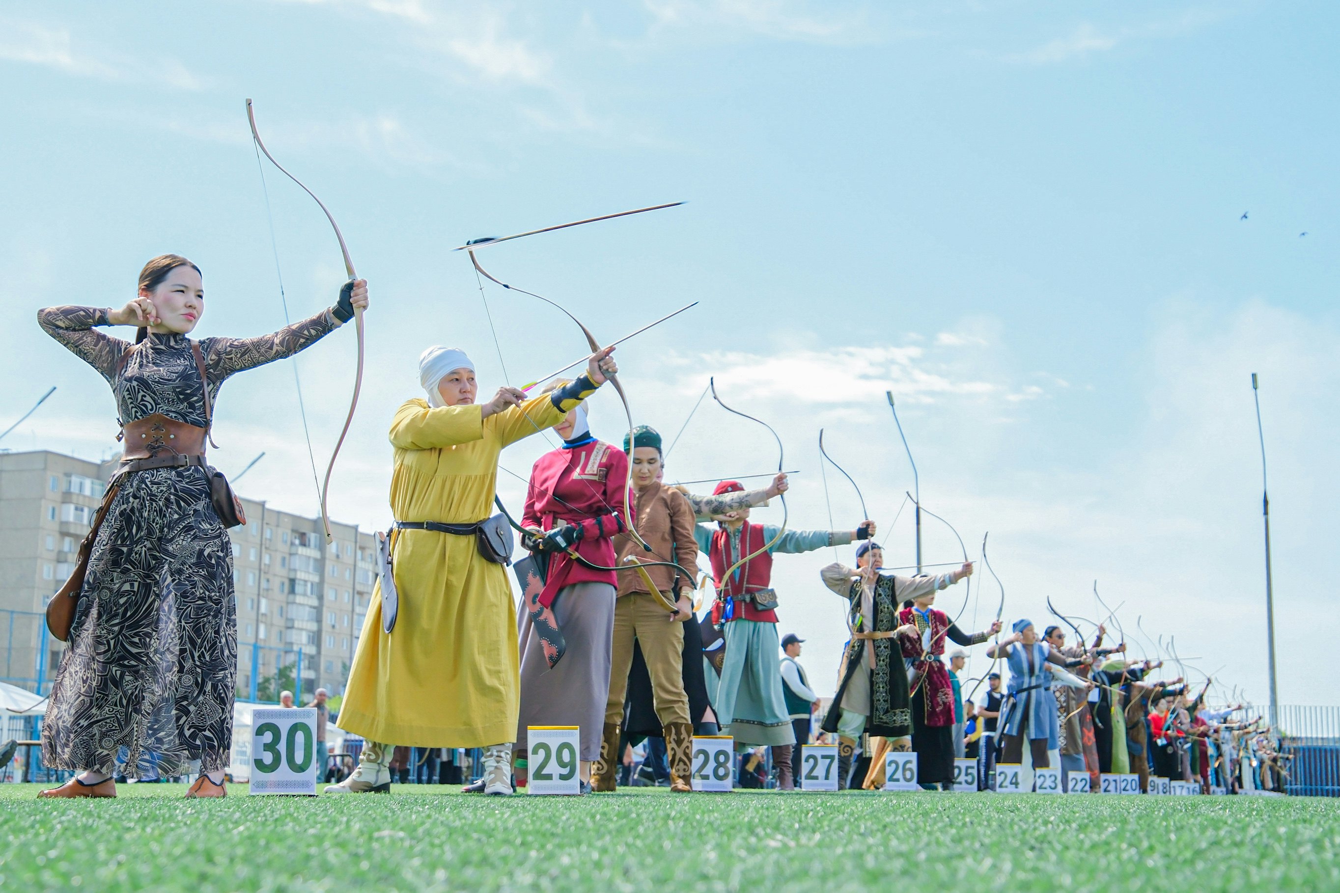 Women in traditional dress of their nations line up with bows and arrows in an archery contest