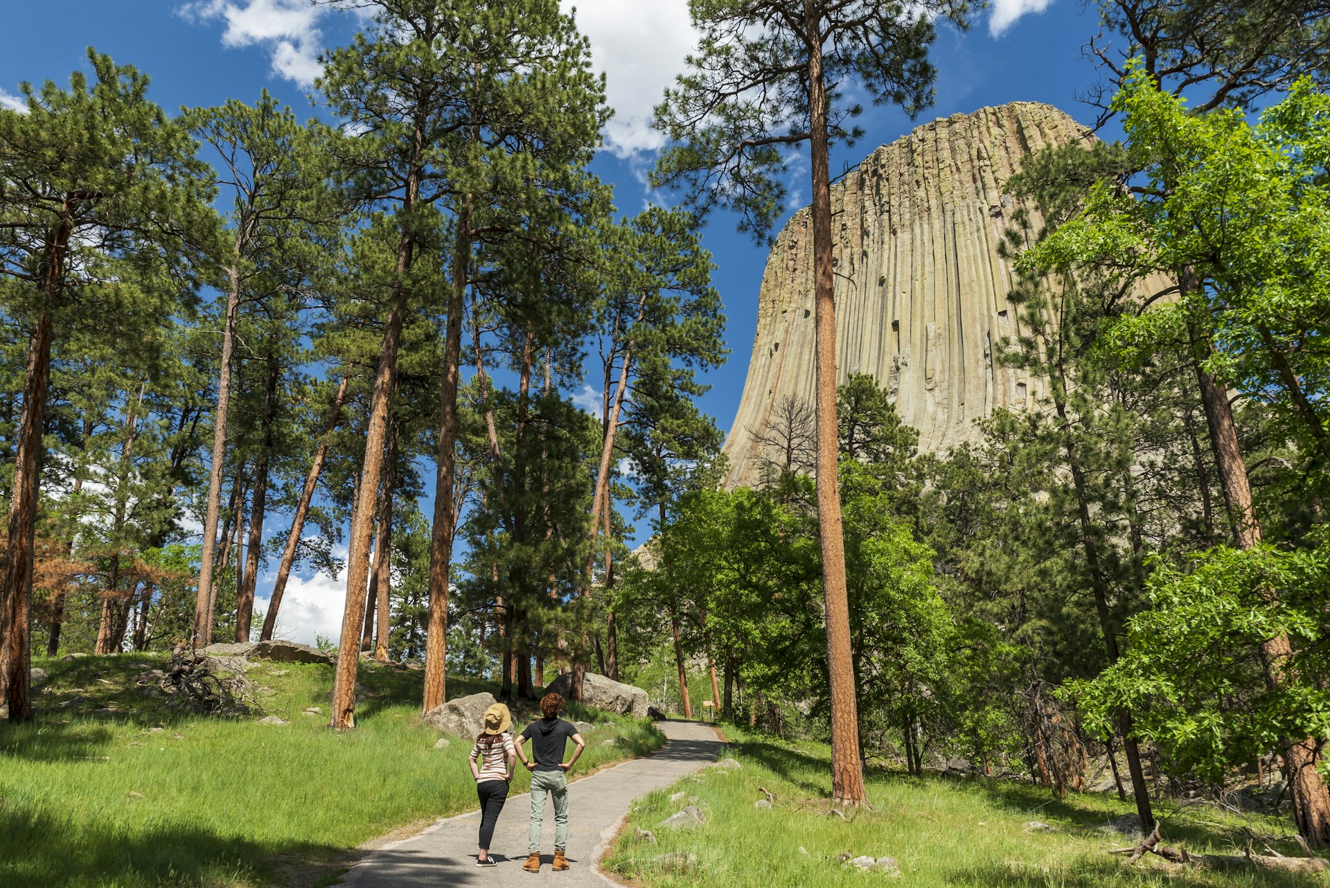 Two hikers are hiking on a trail around Devil Towers, Wyoming, United States. They stop to admire the beautiful forest and the mountain.
