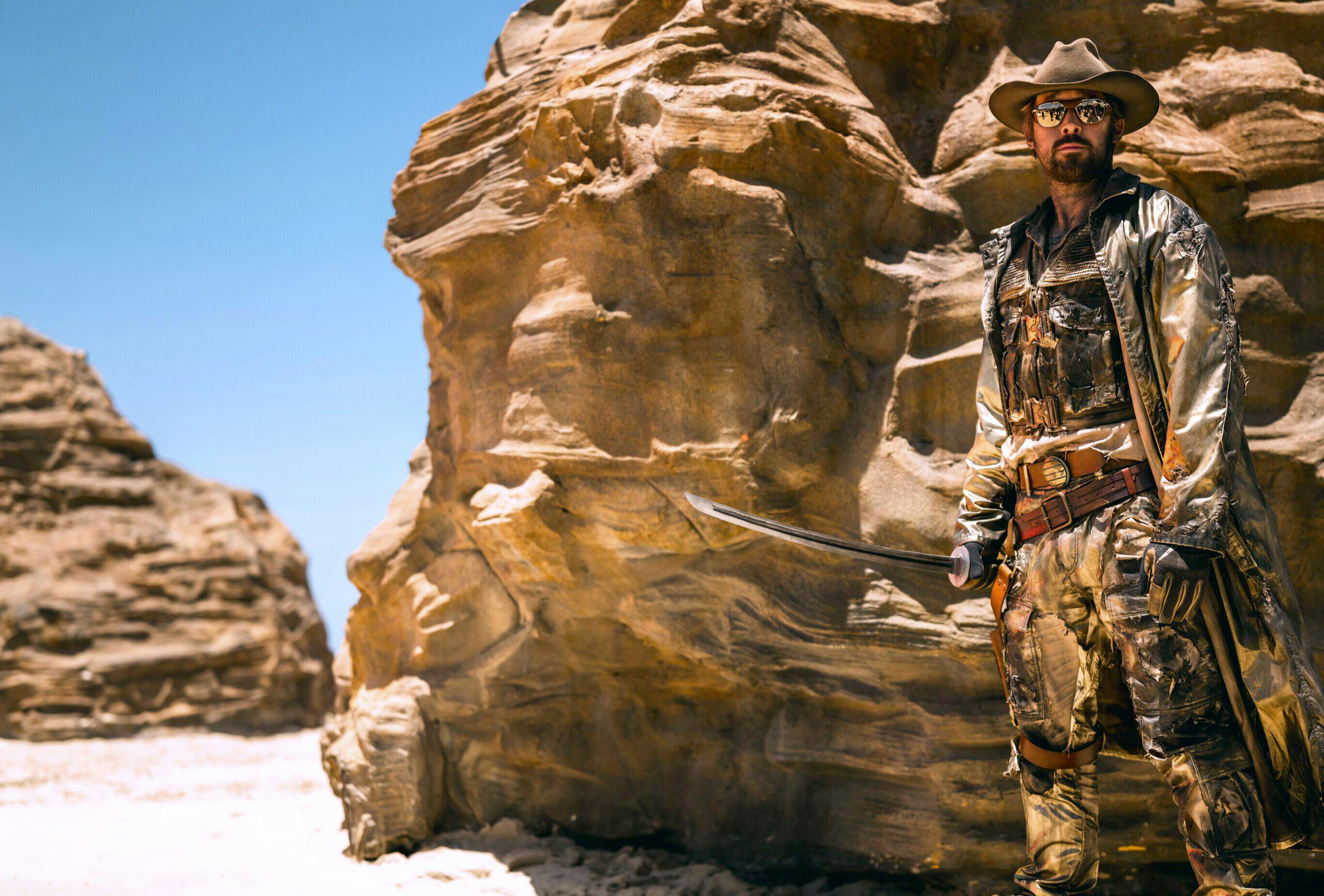 A man stands near a rocky outcrop holding a sword. A group of people are reflected in his sunglasses