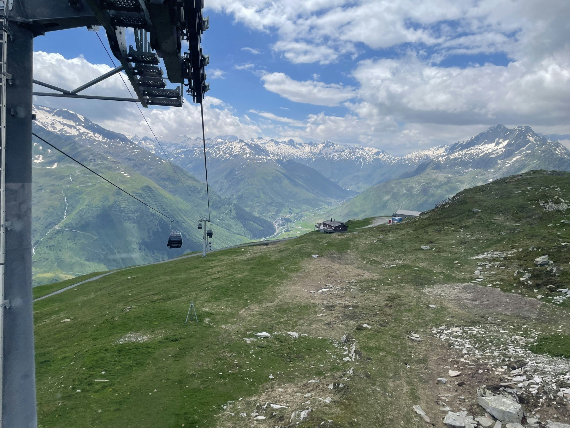 A cable car heads on up a mountain with green rolling hills and snow-capped peaks in all directions