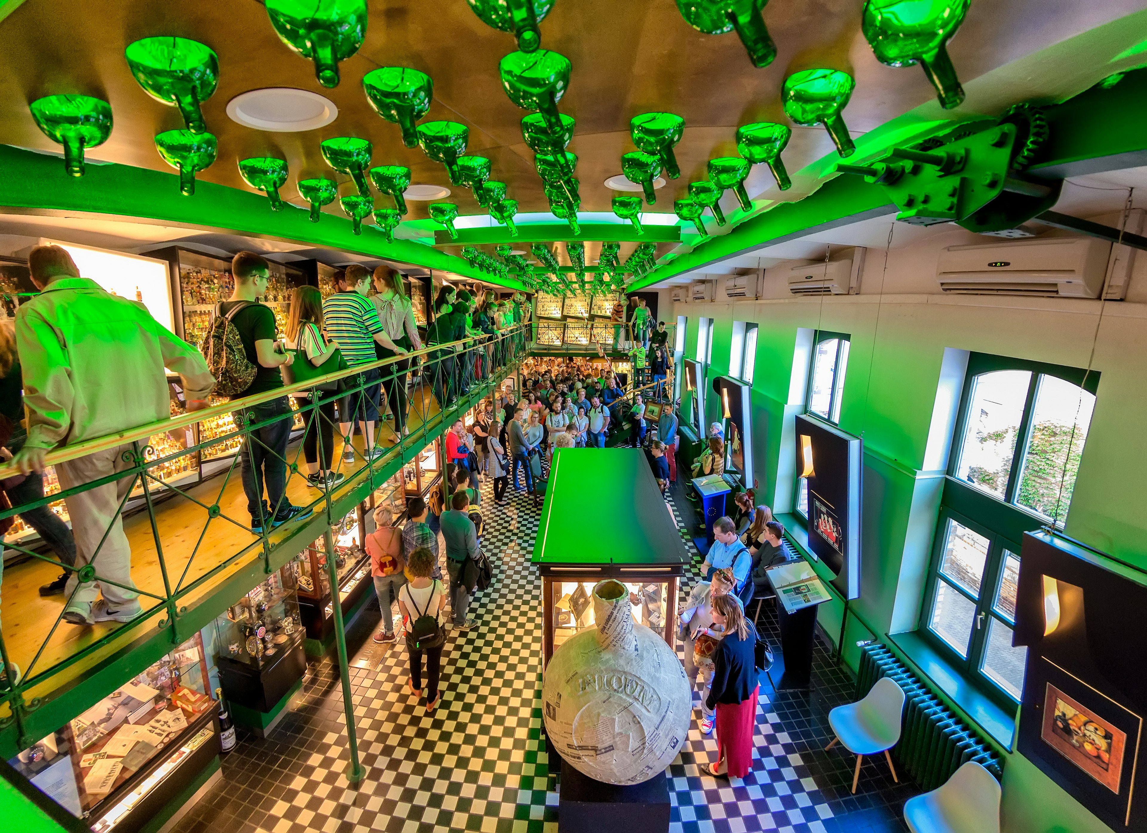 Many people are looking at exhibits over two floors at the Zwack Unicum Factory museum. The exhibition space is decorated with green bottles set into the ceiling.