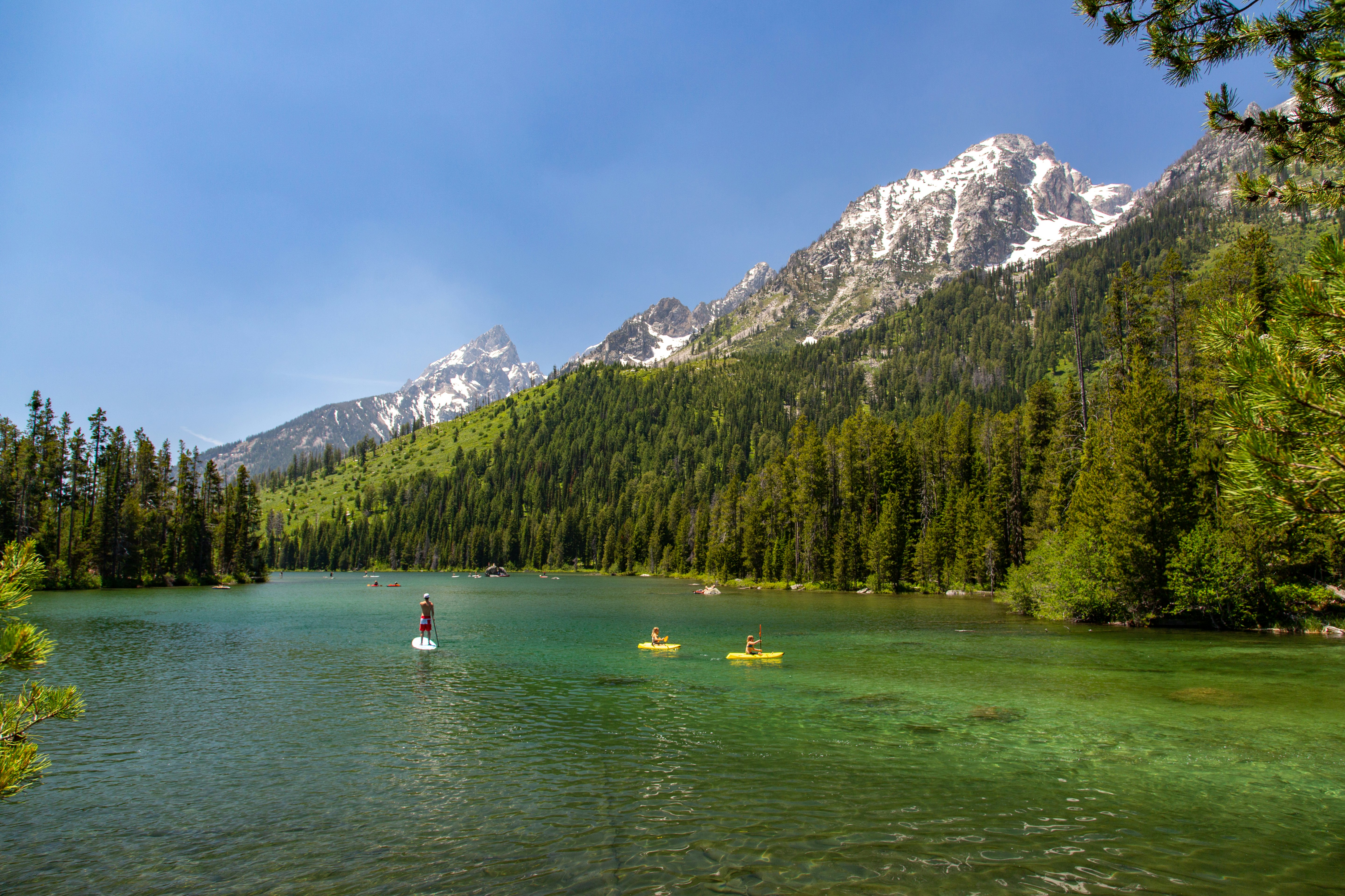 People paddle board and kayak across clear water at the base of a mountain in Grand Teton National Park