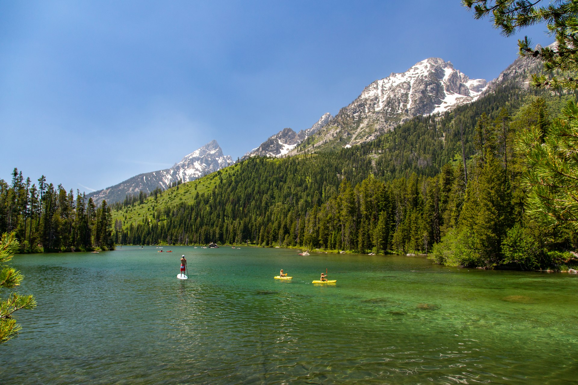 People paddle board and kayak across clear water at the base of a mountain in Grand Teton National Park