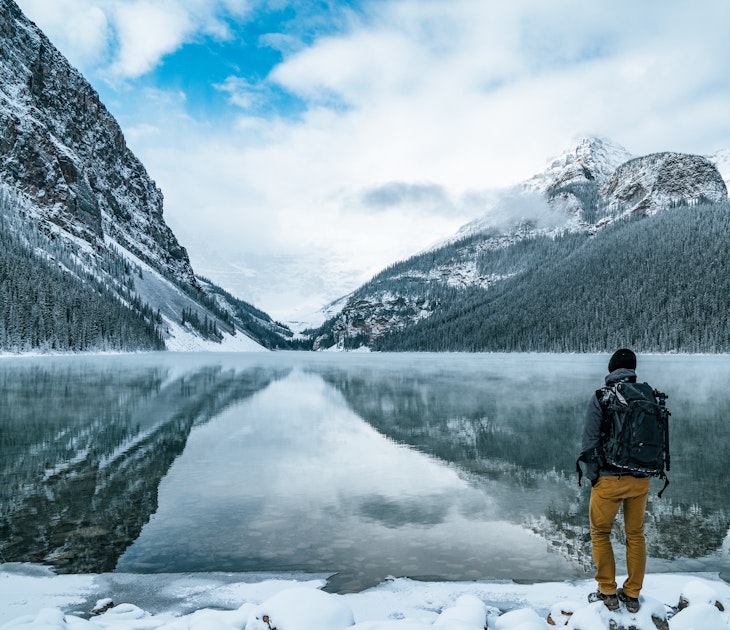 Male hiker enjoys the nature landscape outdoors with backpack at iconic Lake Louise in Banff National Park, Alberta, Canada. Majestic rocky peaks covered with snow reflect on the calm water surface. © Alexander_Magnum / Shutterstock