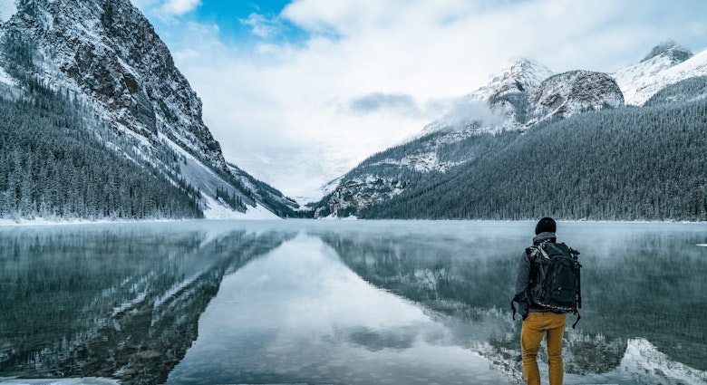 Male hiker enjoys the nature landscape outdoors with backpack at iconic Lake Louise in Banff National Park, Alberta, Canada. Majestic rocky peaks covered with snow reflect on the calm water surface. © Alexander_Magnum / Shutterstock