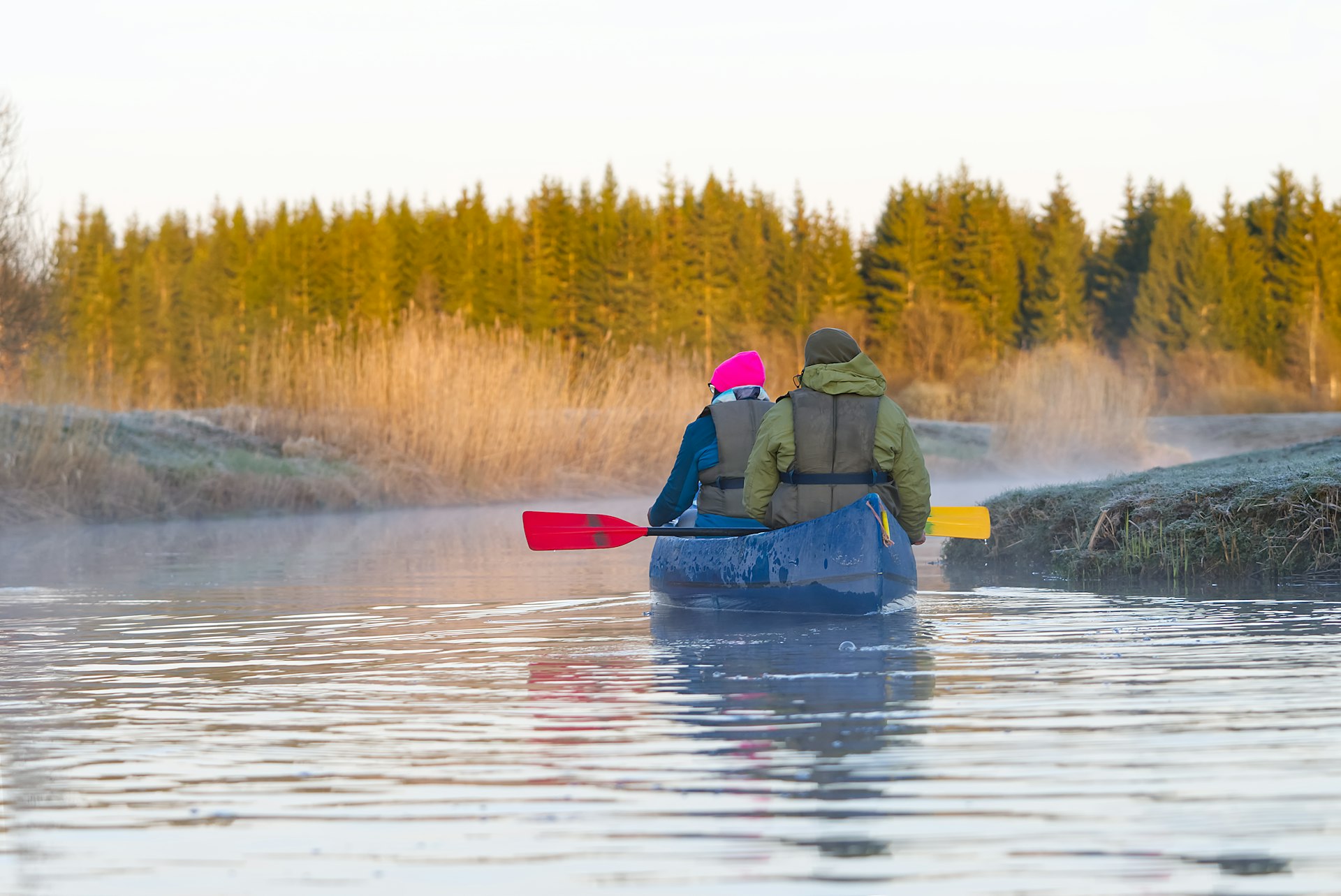 Two people in a kayak paddle their way along a river towards some woodland on a chilly morning