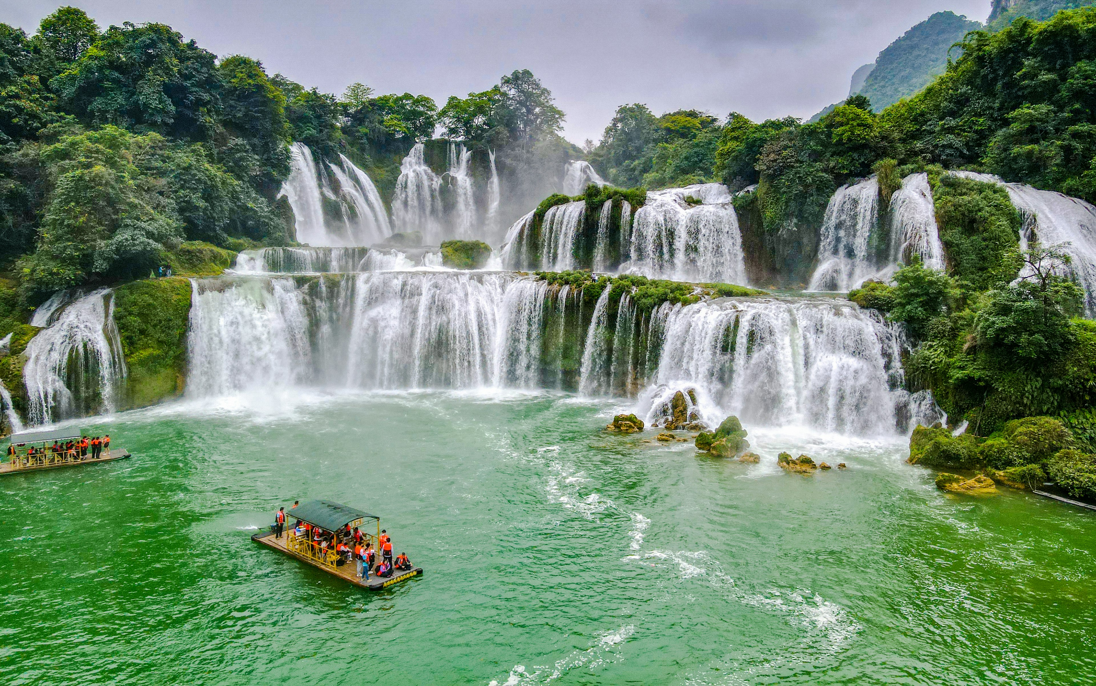 A many-tiered waterfall with a bamboo raft full of tourists in the pool