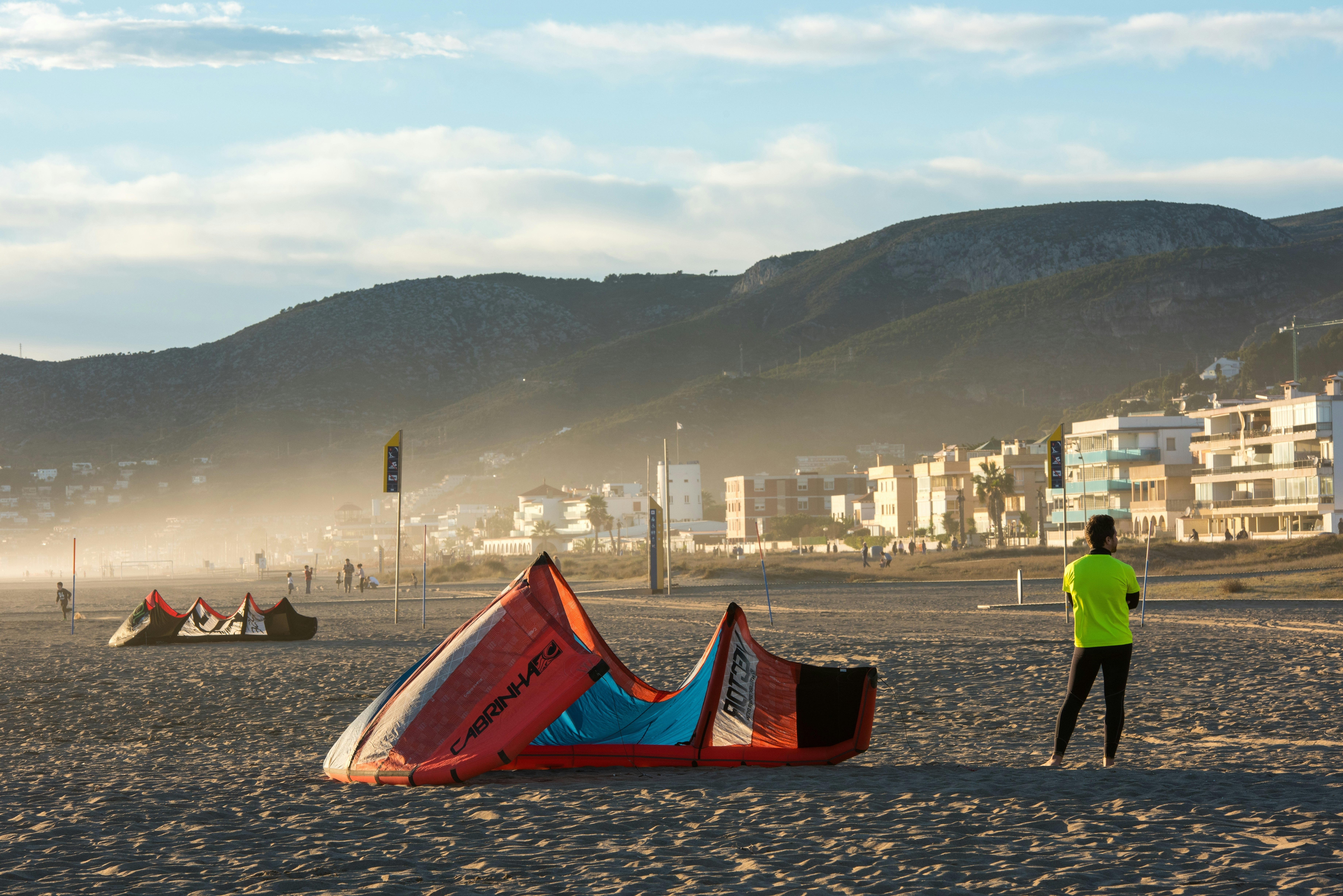 Young man with neoprene and lycra shirt waits next to his kite on the sand of the beach, with houses of the promenade in the background, Castelldefels, Catalonia, Spain