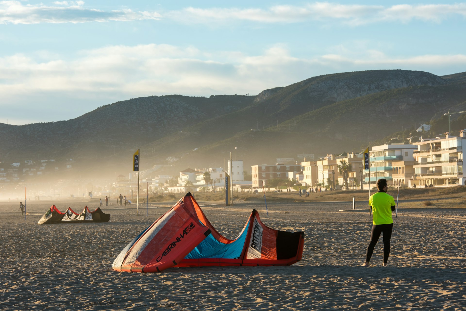 Young man with neoprene and lycra shirt waits next to his kite on the sand of the beach, with houses of the promenade in the background, Castelldefels, Catalonia, Spain