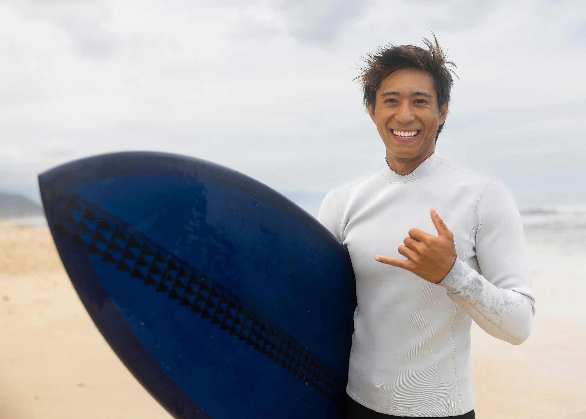 A smiling man on a beach is holding a surfboard while making the "shaka"hand gesture