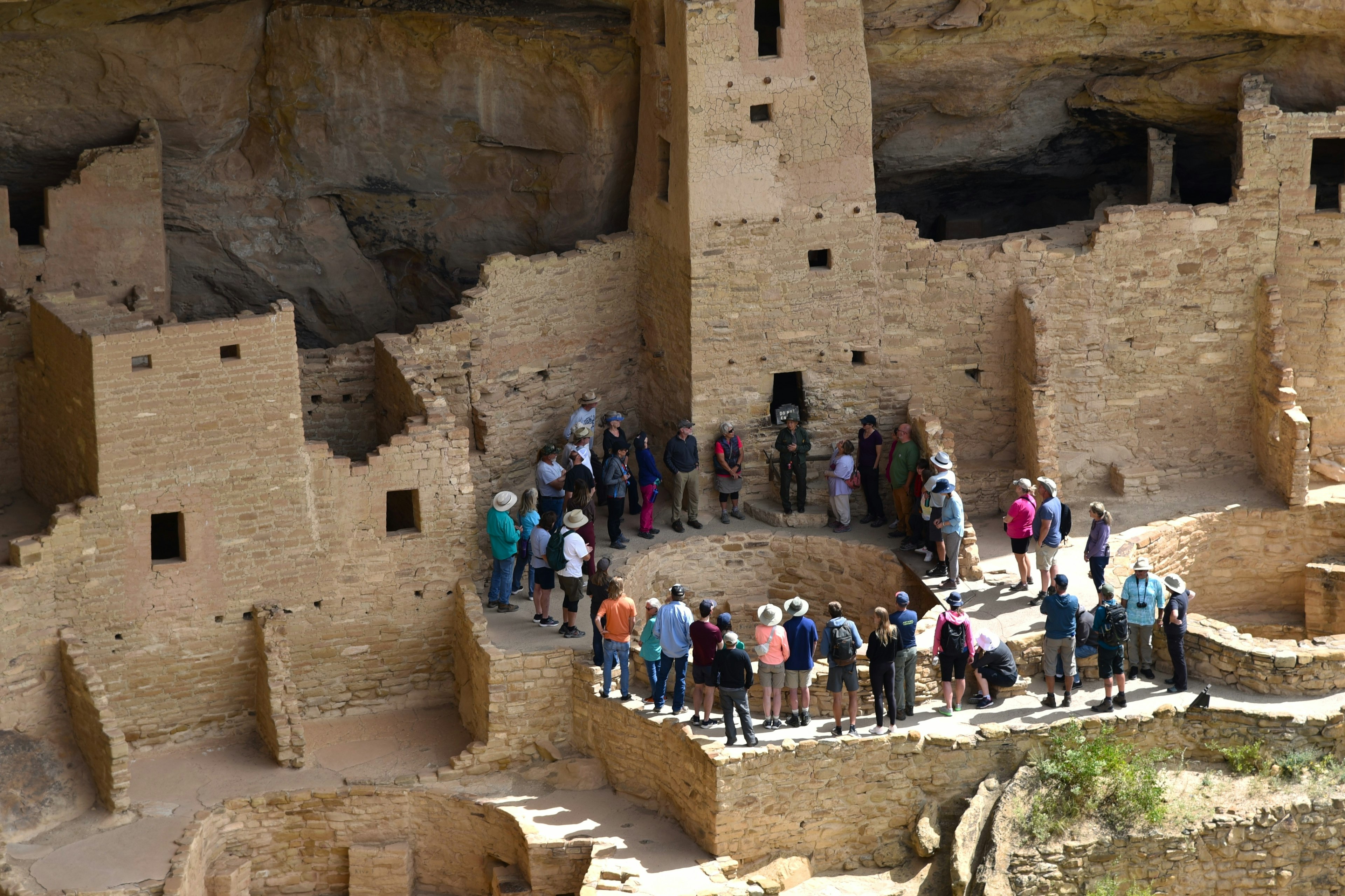 A group of tourists stand in a circle in an ancient site built into the side of a cliff as they listen to a guide