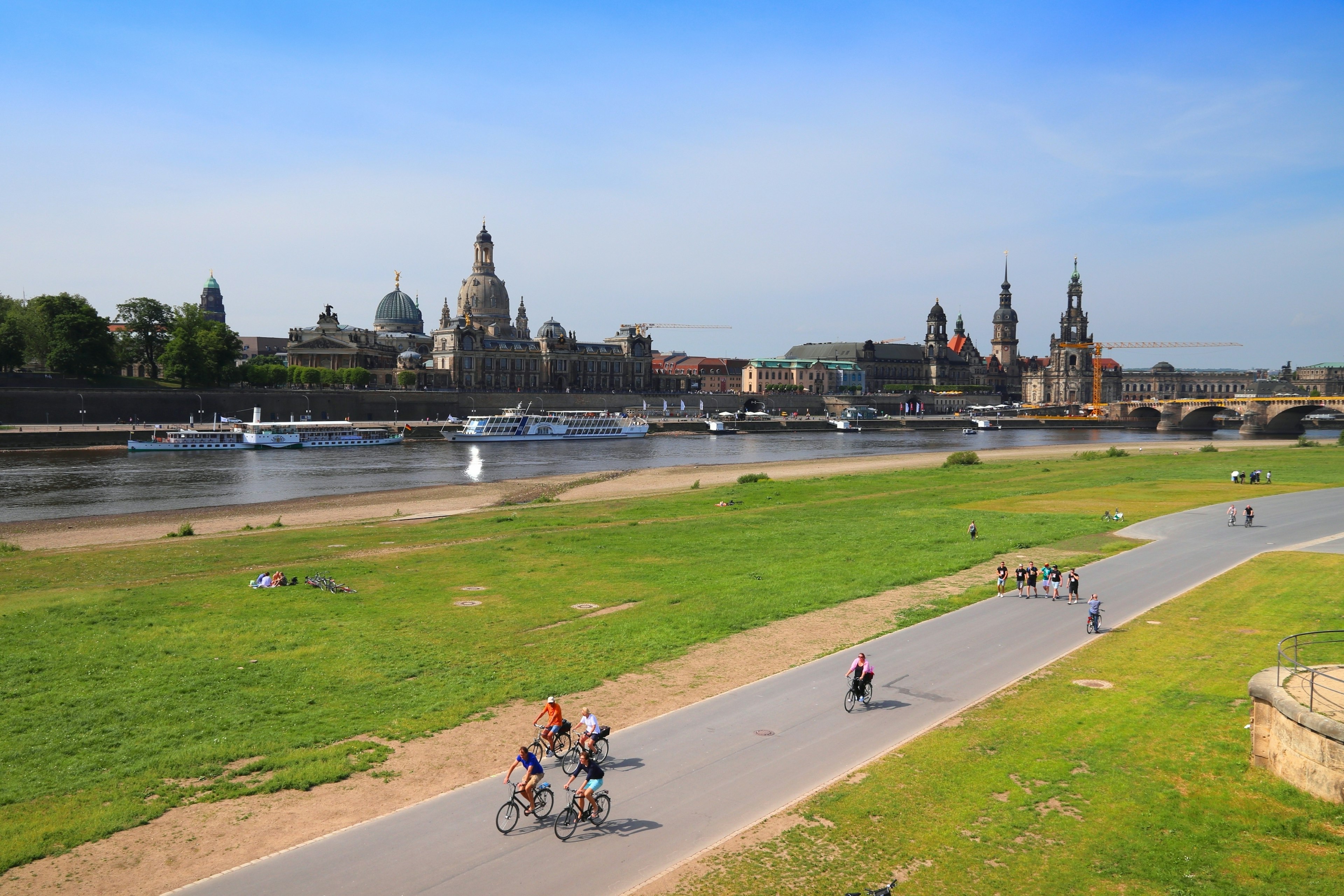 Cyclists ride on a path beside a river. A city with spires and domes is across the river