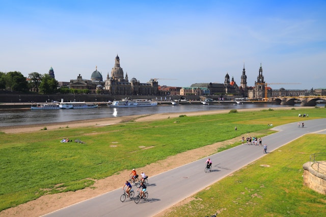 Cyclists ride on a path beside a river. A city with spires and domes is across the river