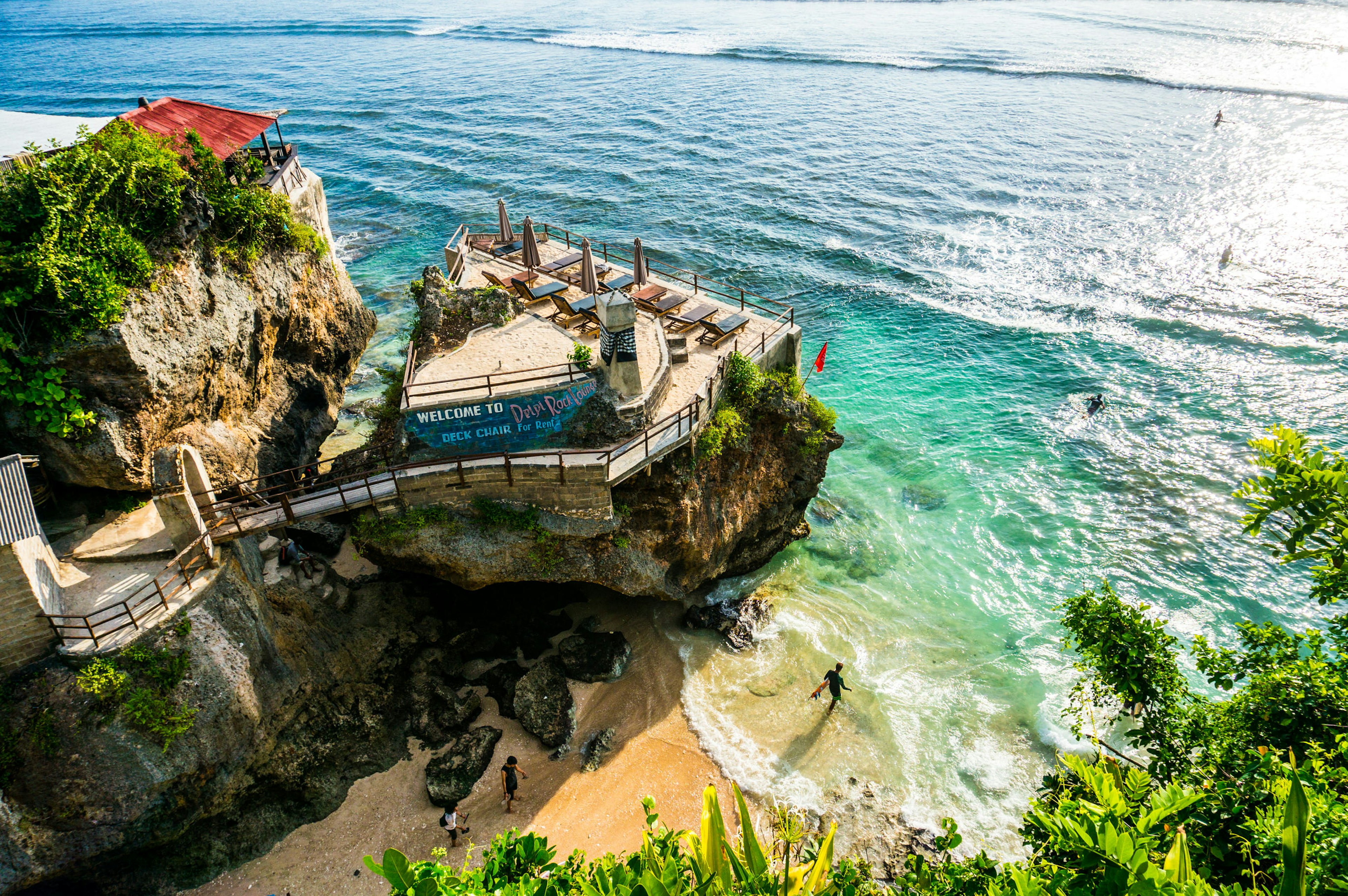 A surfer walking out to the ocean near a sheer cliff with a bar-lounge on top