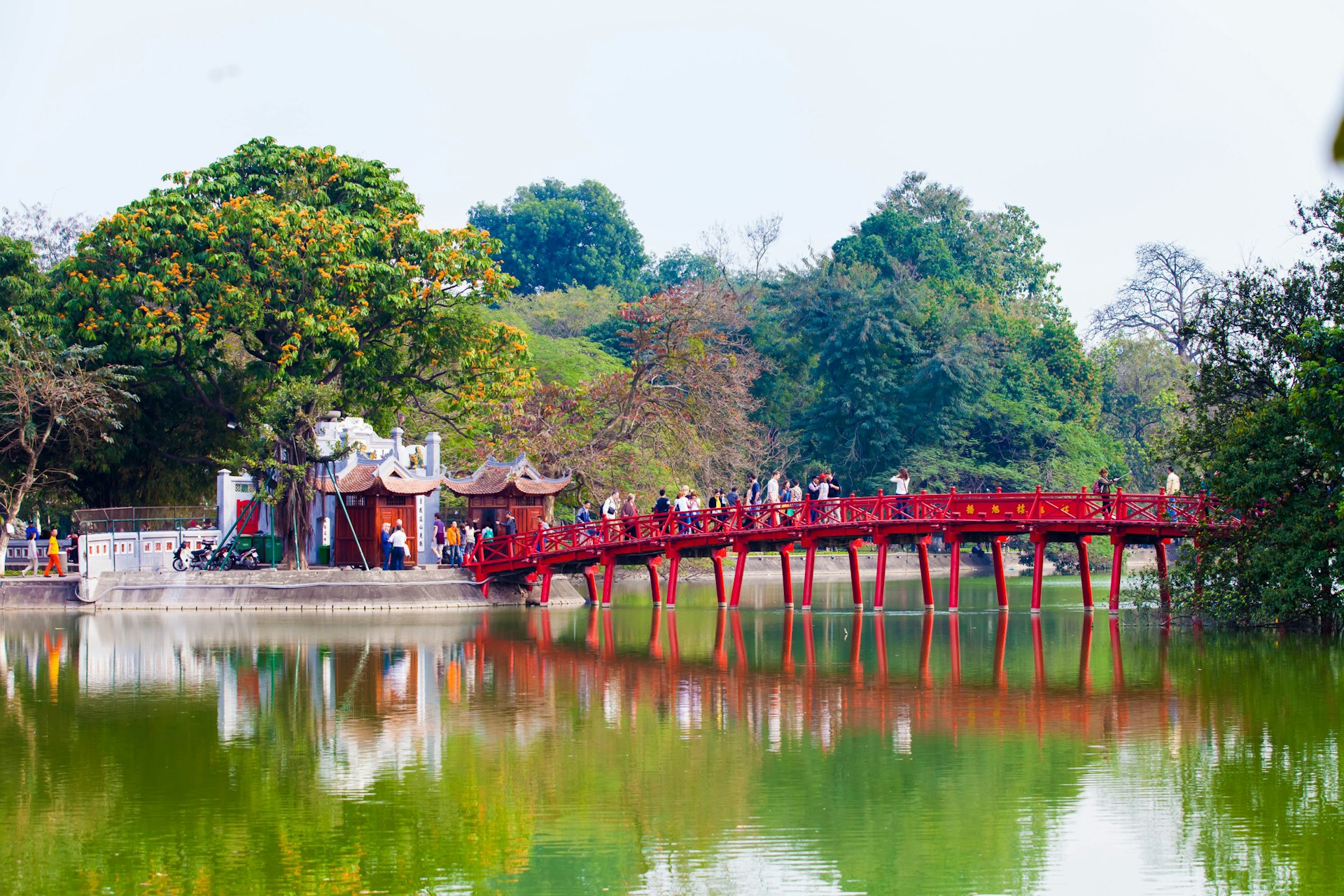 People walk over a red bridge leading to a temple on a lake