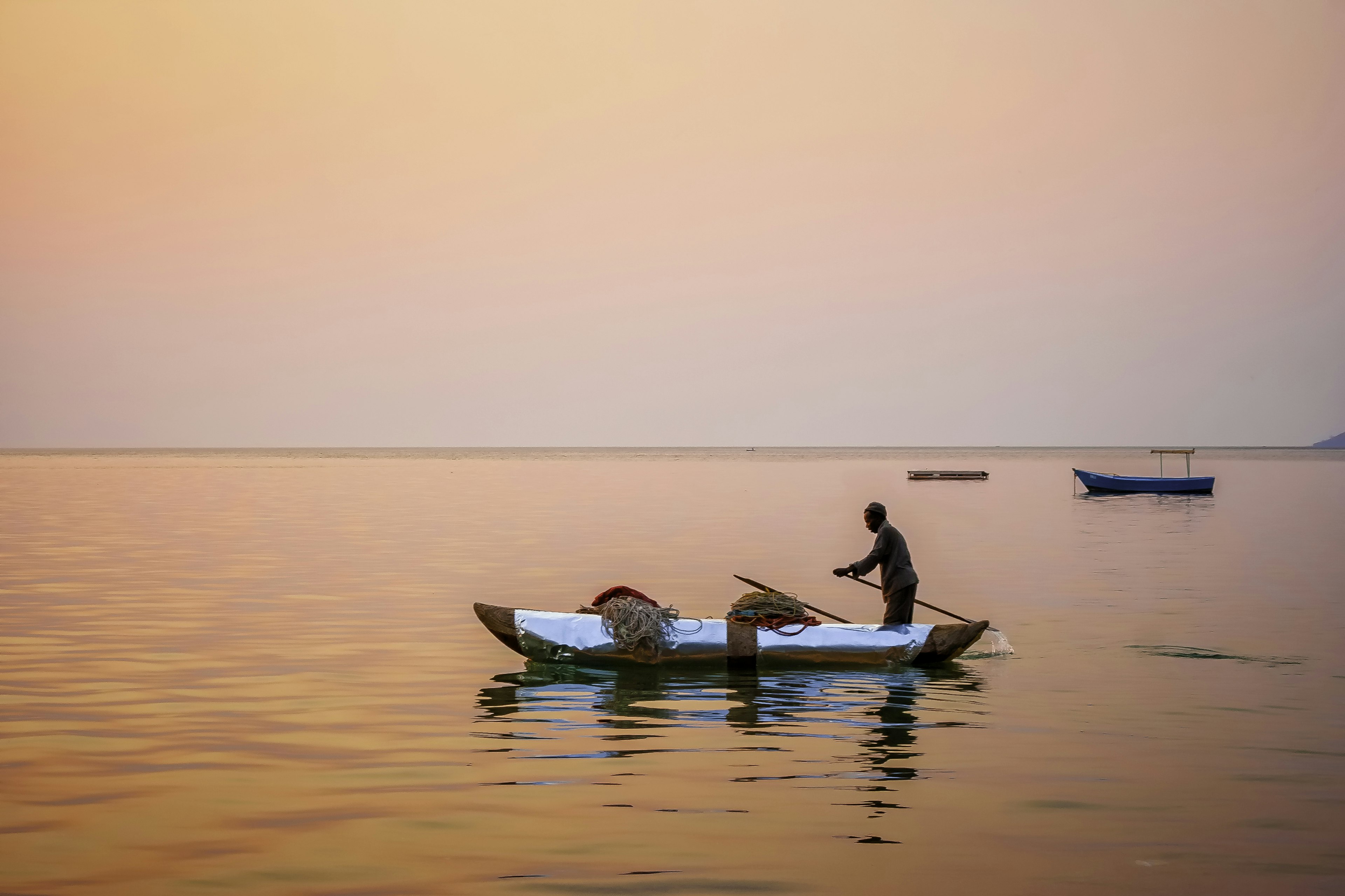 People in silhouette fishing from a canoe at sunset, with an orangey-pink sky reflected in the water.