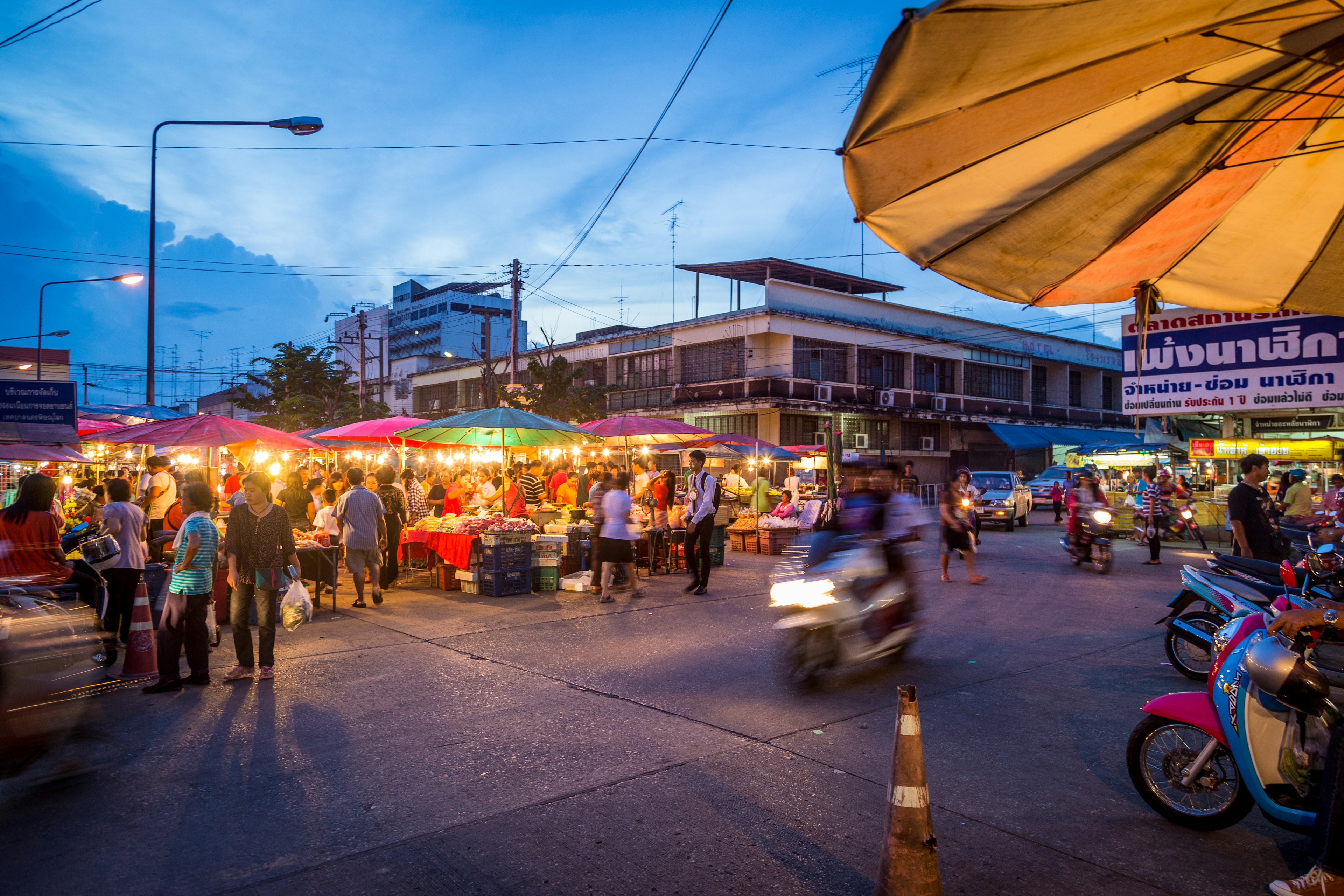 A blurred-out motorcycle drives past crowds at a night market on the streets of Phitsanulok, Thailand at dusk