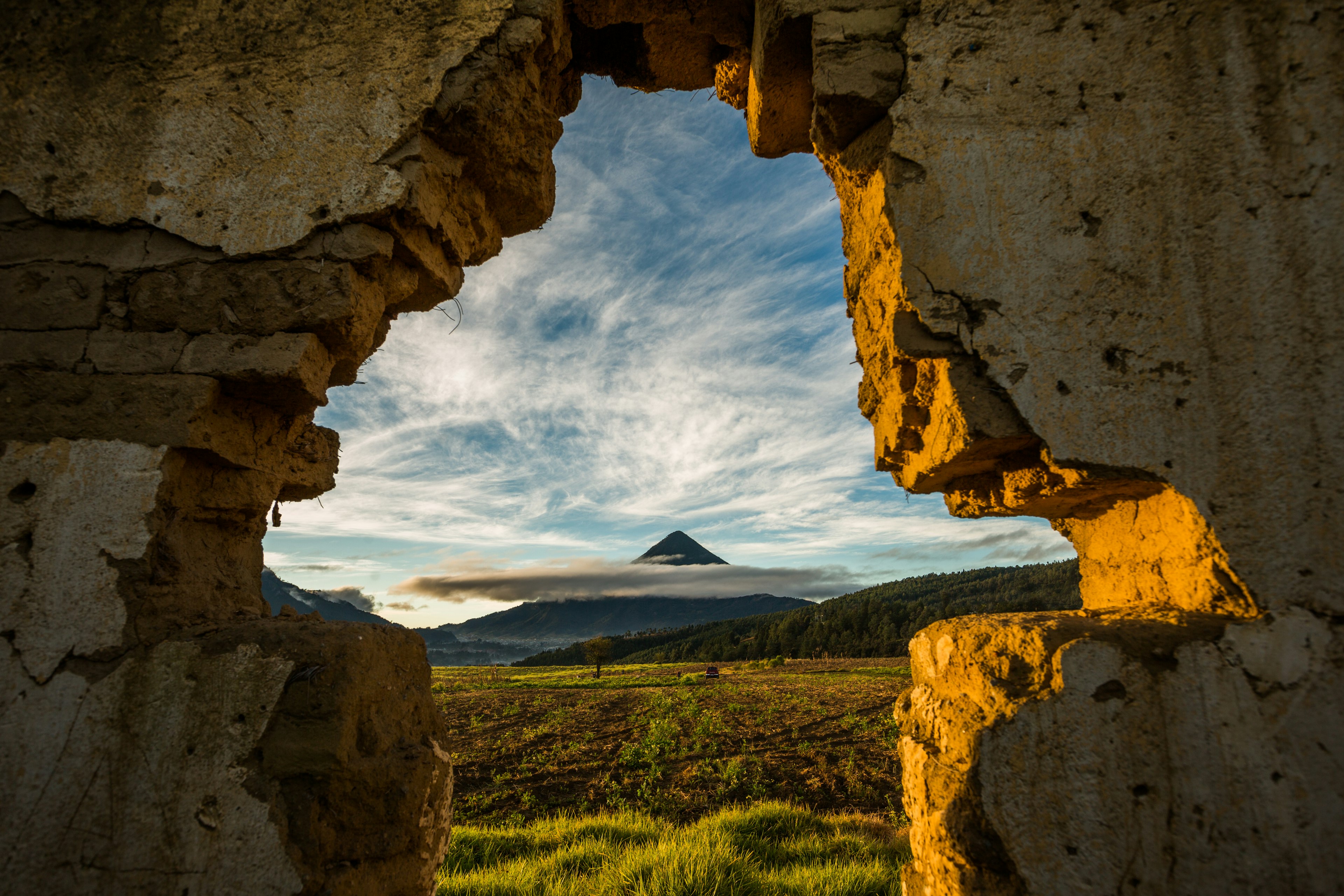 A cone-shaped volcano topped with clouds, framed by a broken stone wall in Quetzaltenango, Guatemala.