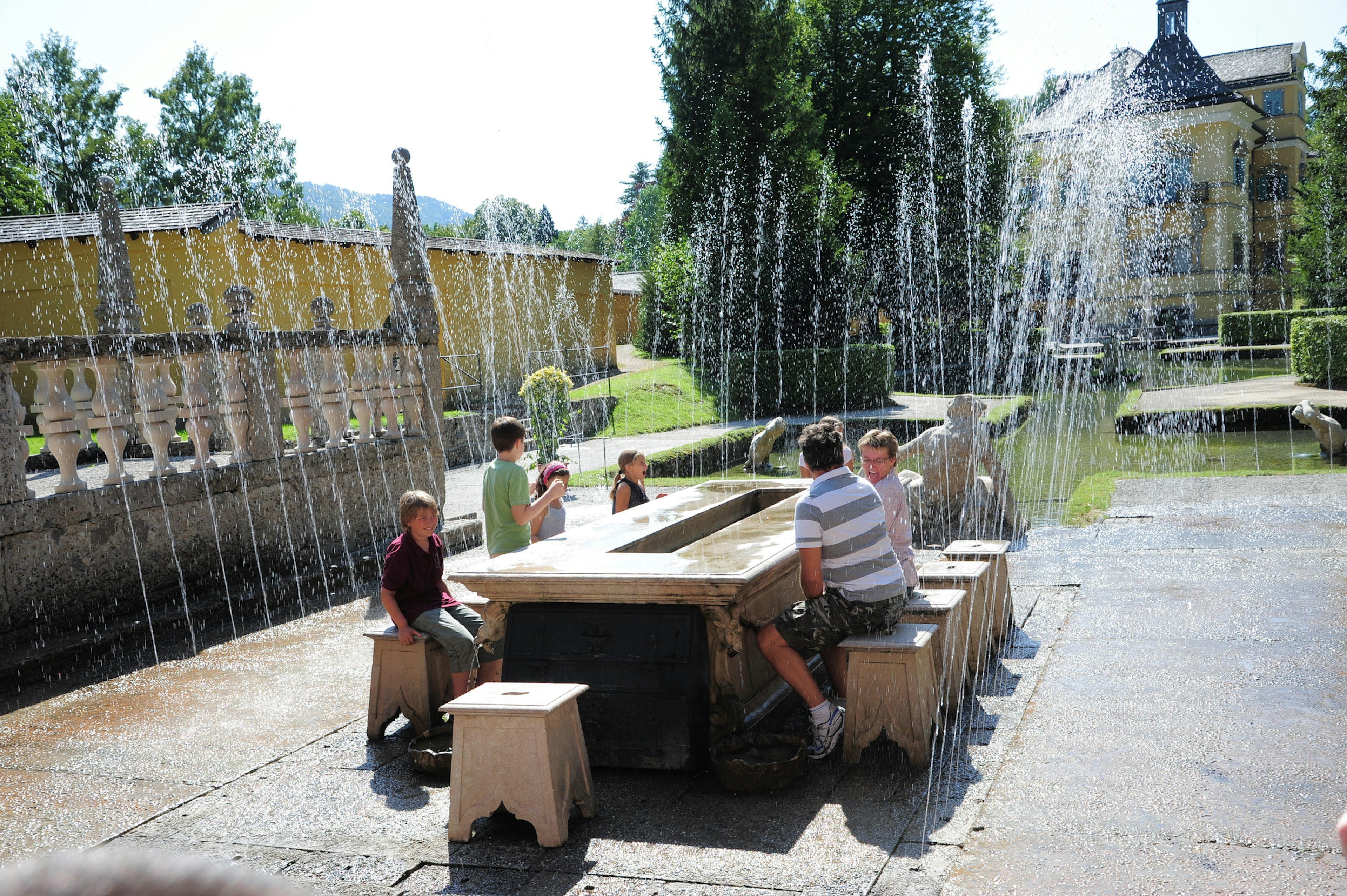 A family sit on ornate stone chairs around a table outdoors in Hellbrunn Palace Park as surprise fountains go off around them