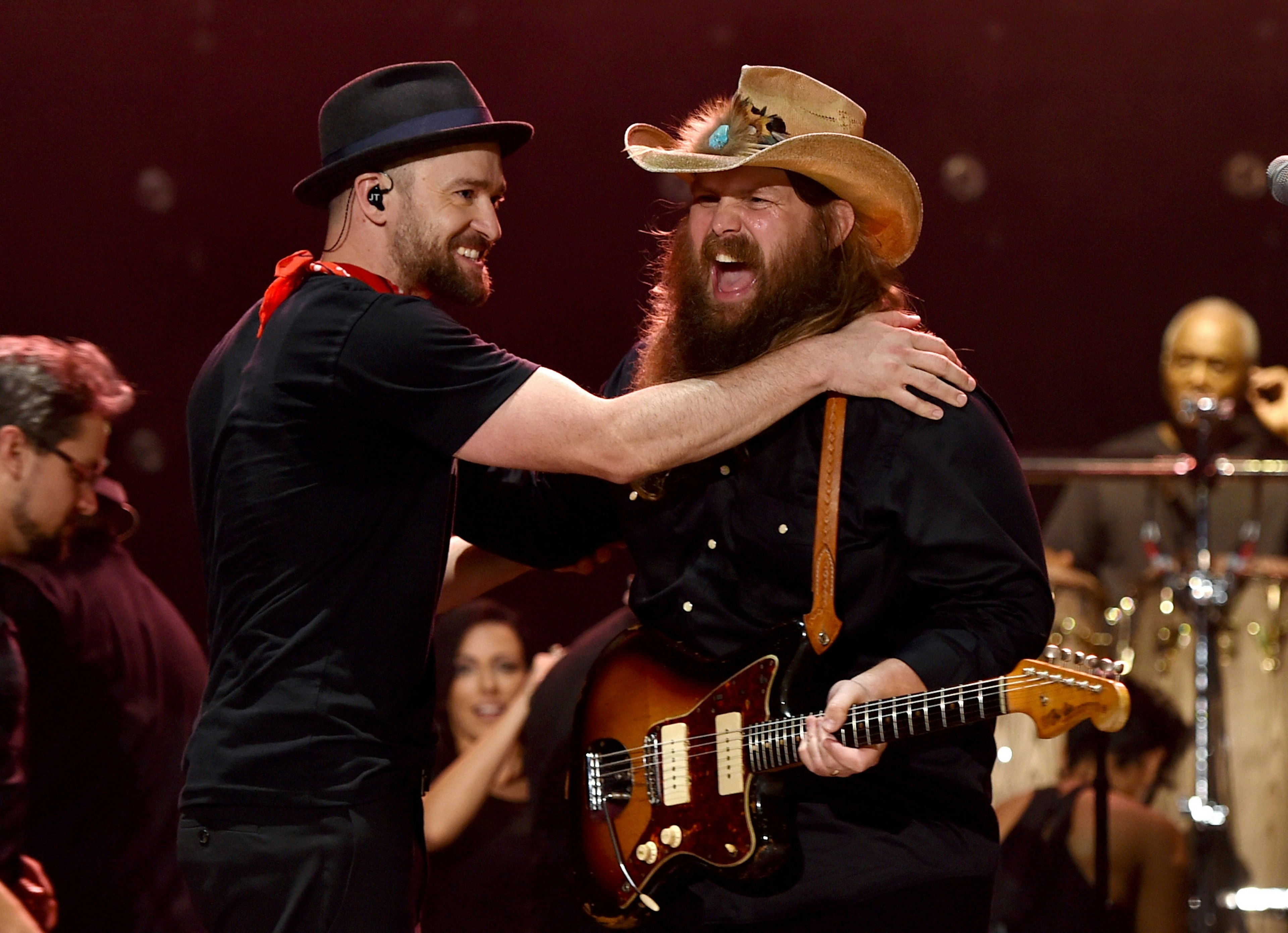 Two men perform on stage; the one on the left is patting the other man on the shoulder while he plays guitar