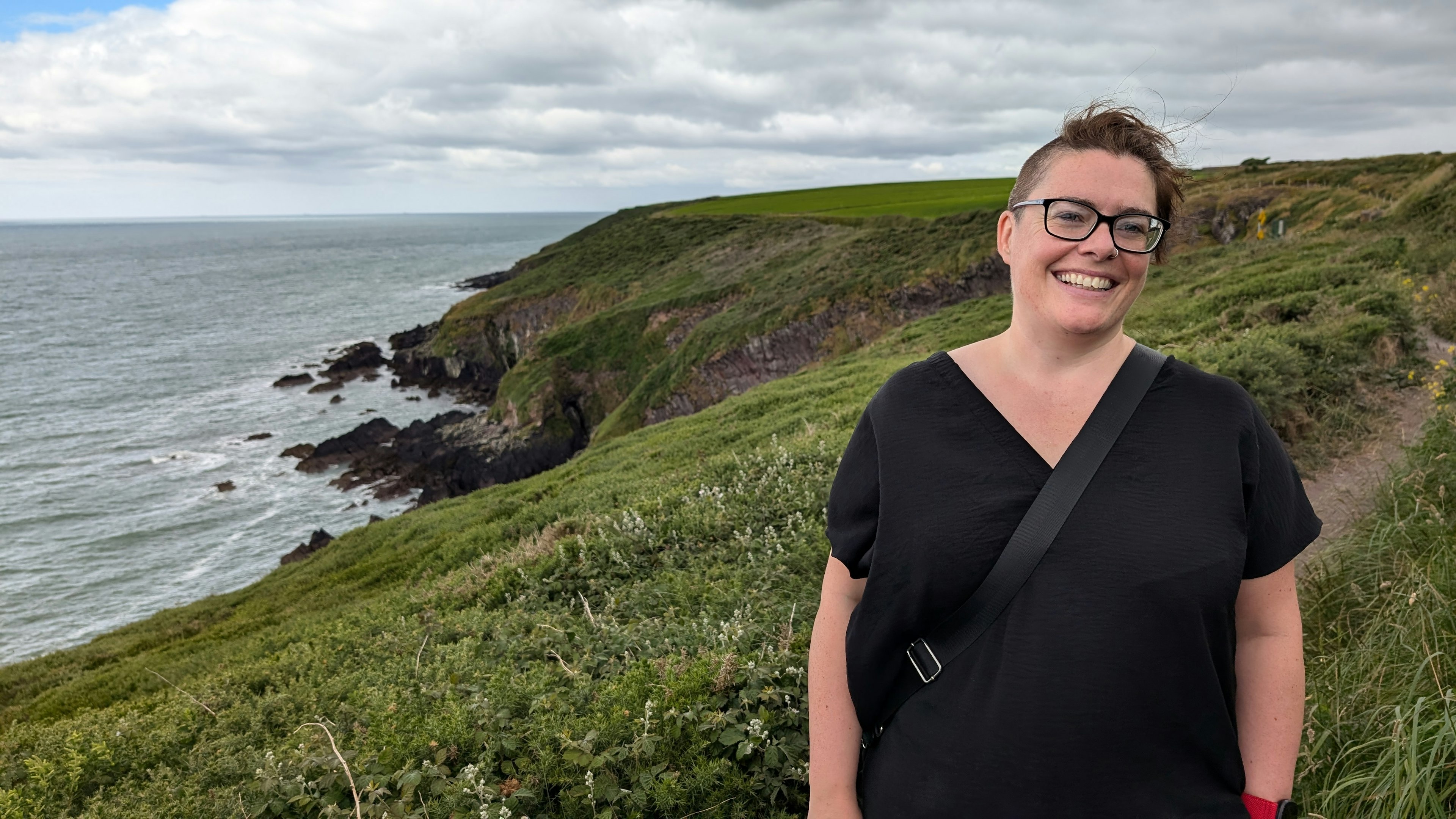 A portrait of author Amy Lynch along the grassy trail of the Ballycotton Cliff Walk with the sea in the distance