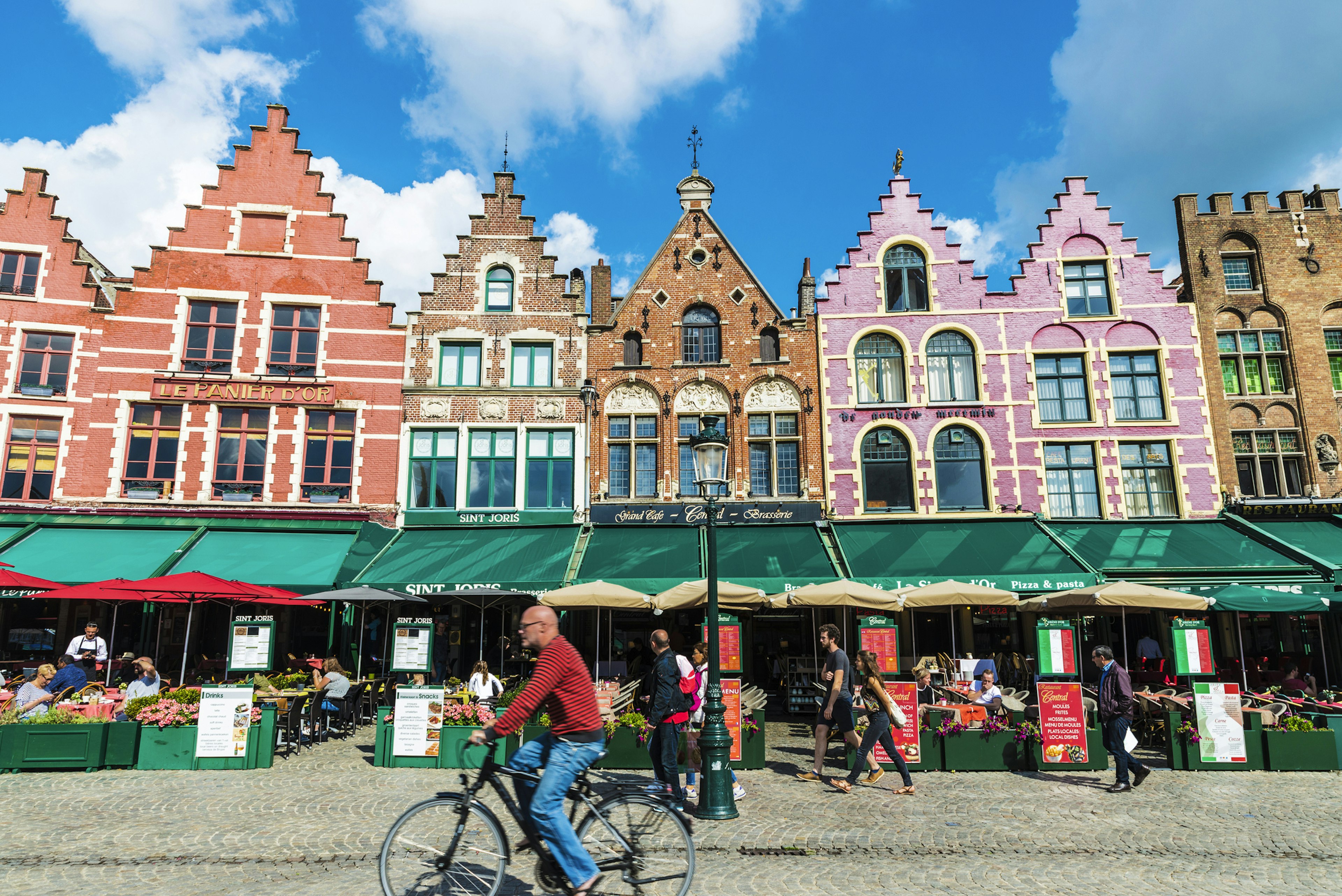 Pastel-colored tall buildings line a square with cafes and restaurants at the base. A cyclist whizzes by.